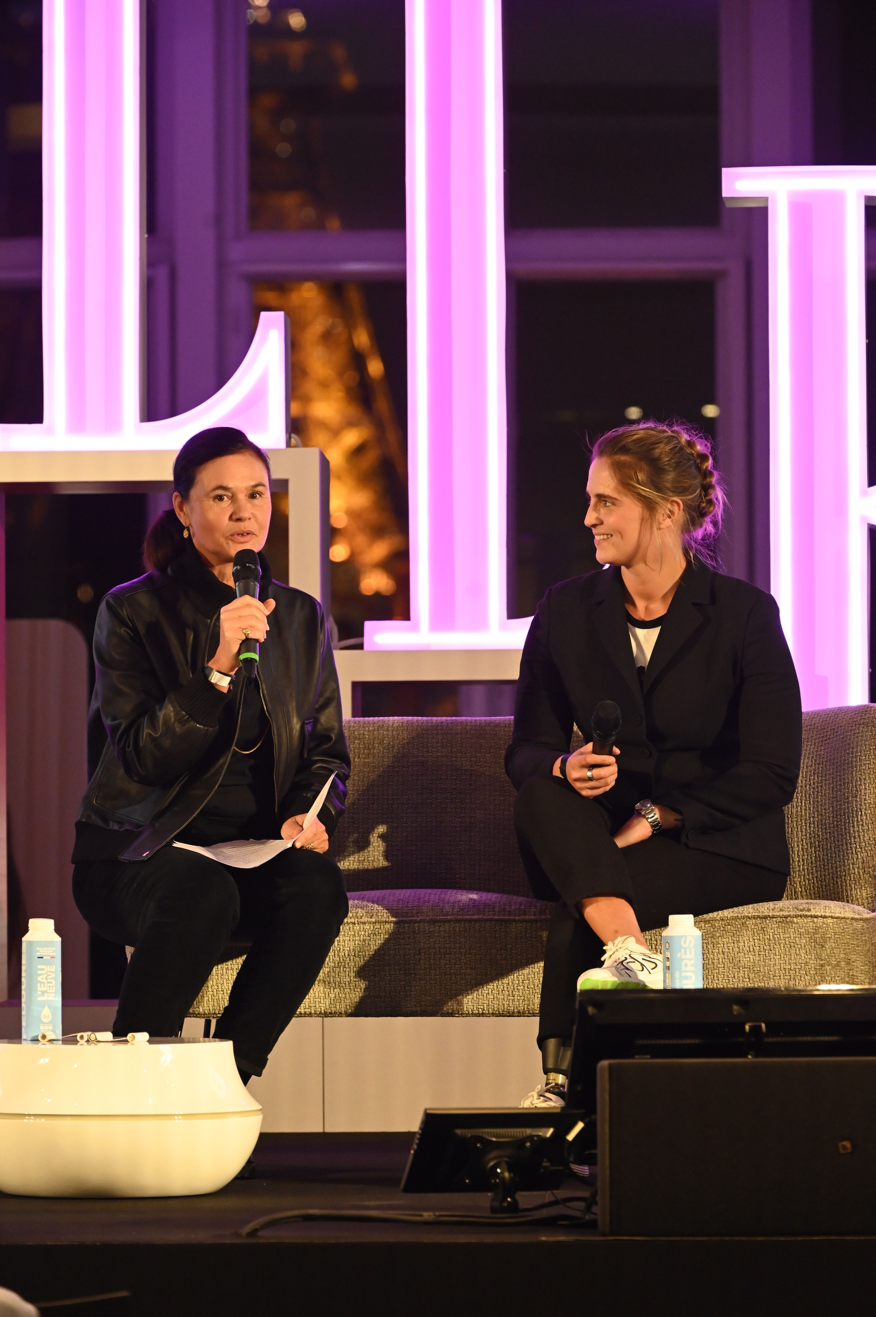 Deux femmes sont assises sur un canapé sur une scène éclairée de lumières violettes, tenant chacune un micro. L'une d'elles, vêtue d'une veste en cuir noir, s'adresse au public en lisant un document. L'autre, en costume noir et baskets blanches, l’écoute avec le sourire. En arrière-plan, on distingue une grande structure lumineuse formant les lettres "ELLE" et une vue floue de la tour Eiffel à travers une baie vitrée. Une ambiance professionnelle et bienveillante se dégage de la scène.