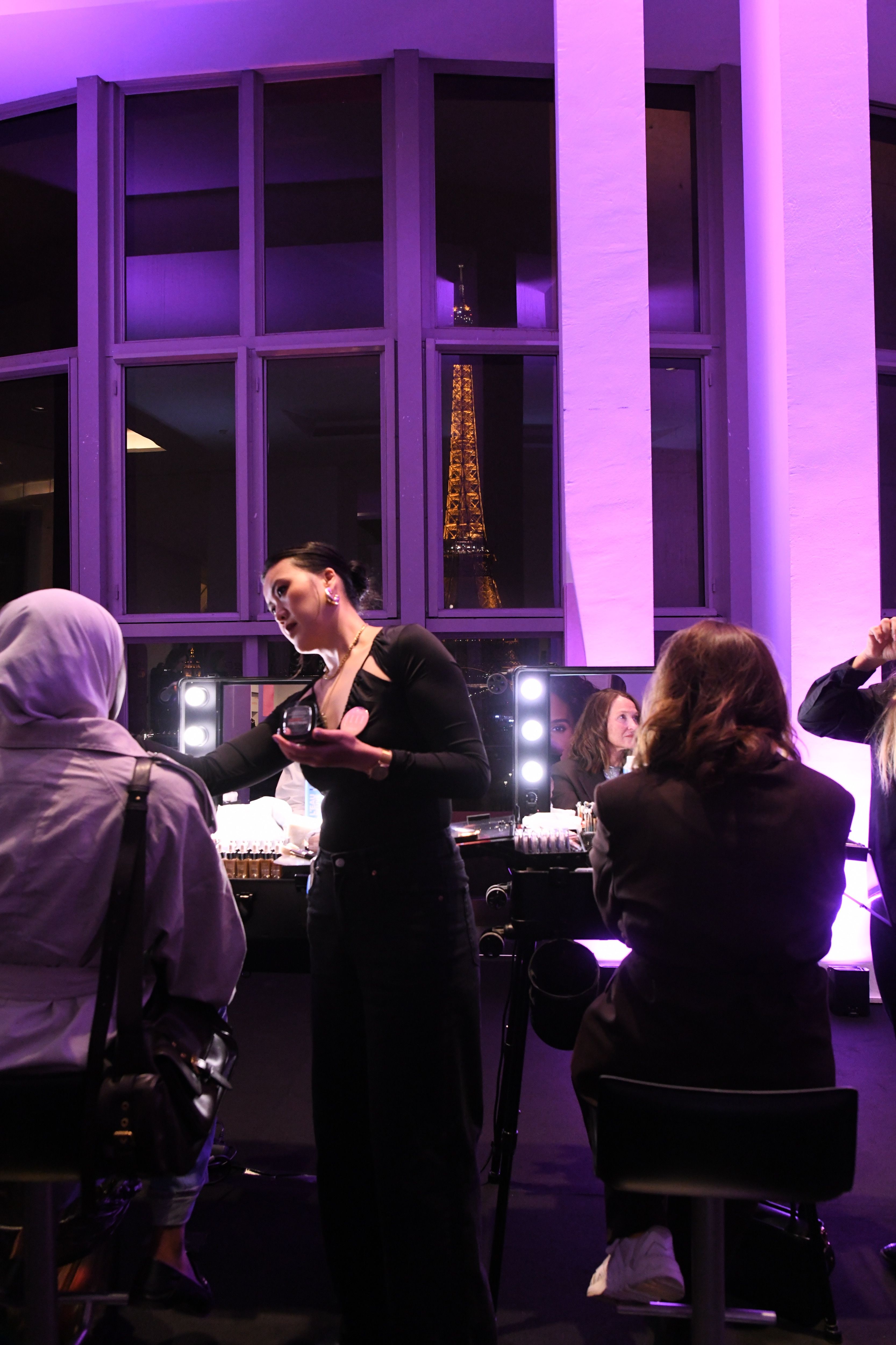 Dans une ambiance tamisée aux tons violets, une maquilleuse professionnelle applique du maquillage sur une femme portant un hijab, assise devant une coiffeuse éclairée par des lumières LED. D'autres femmes sont également installées au poste de maquillage. À l’arrière-plan, les fenêtres révèlent une vue nocturne spectaculaire sur la Tour Eiffel illuminée. L’ensemble crée une atmosphère chic et intimiste, typique d’un événement beauté ou mode à Paris.