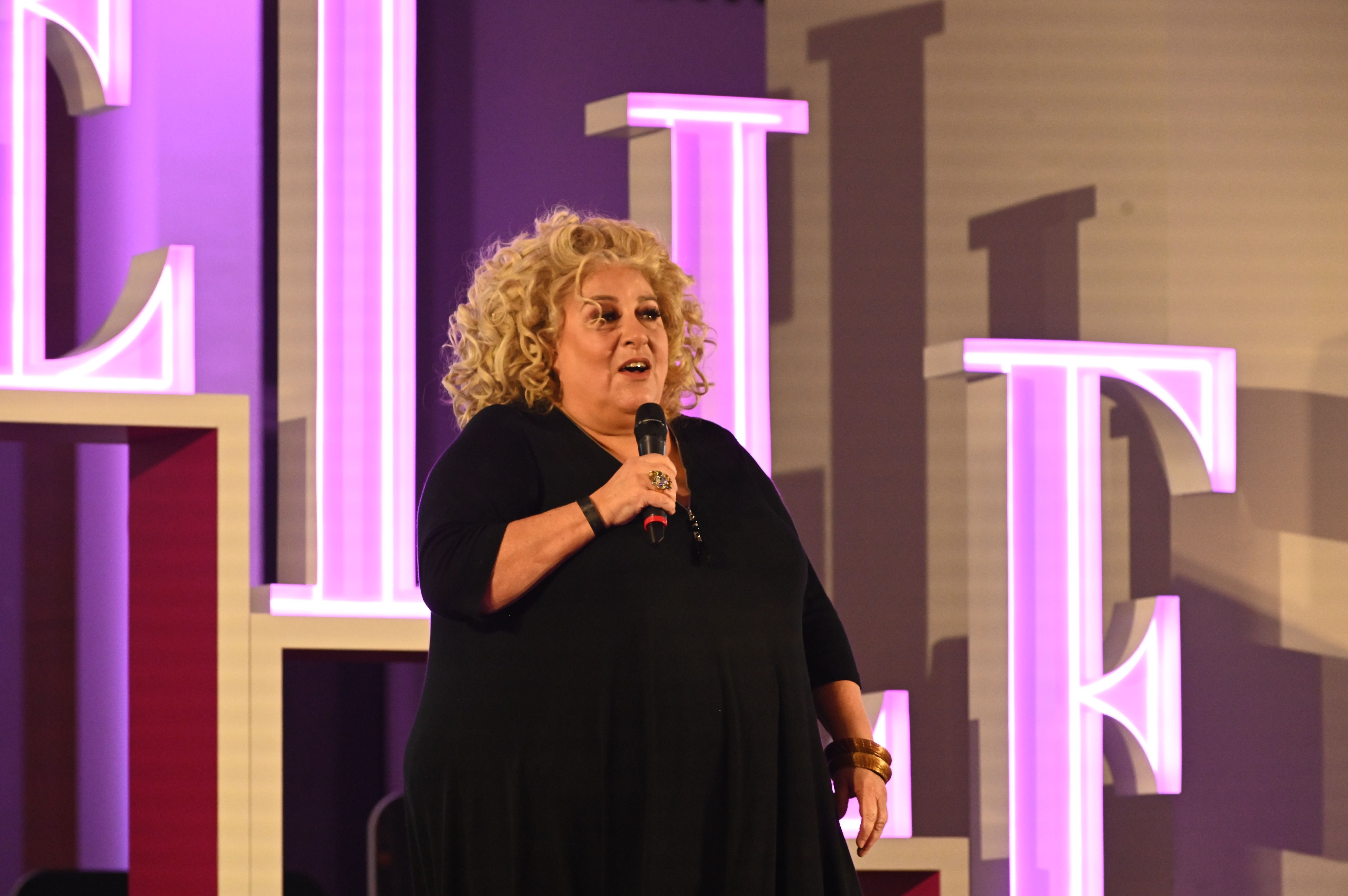 Une femme aux cheveux blonds bouclés, vêtue d’une robe noire, prend la parole sur scène en tenant un micro. Elle est éclairée par des projecteurs et se tient devant des lettres lumineuses formant le mot "ELLE", aux tons violets. L’ambiance suggère un événement ou une conférence organisée par le magazine ELLE.