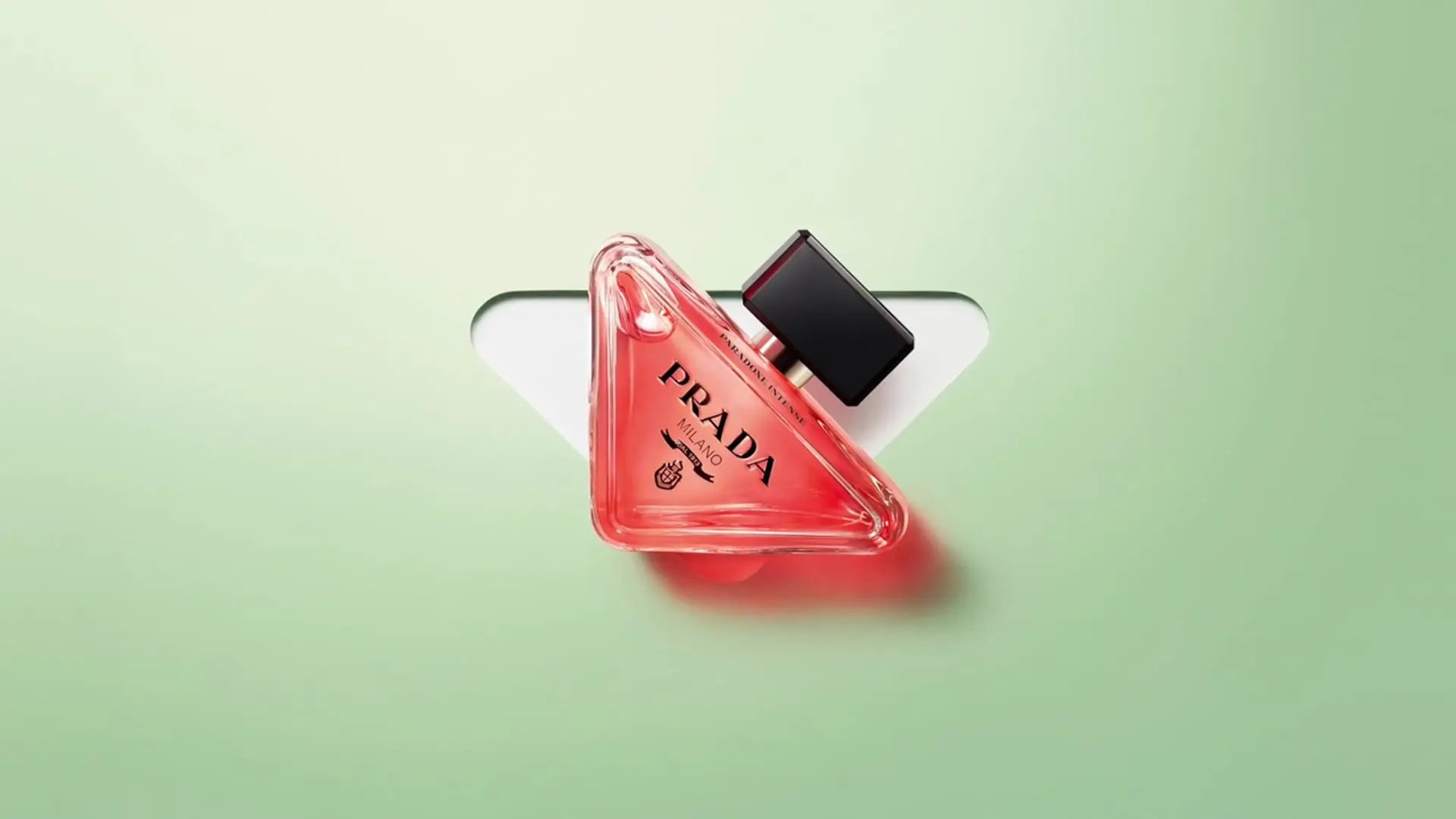 Flacon de parfum en forme de triangle rose translucide avec l'inscription « PRADA MILANO » en lettres noires. Le flacon est incliné et repose contre un fond vert pâle, avec un élément blanc en arrière-plan formant un V stylisé. Le bouchon du flacon est noir et rectangulaire, créant un contraste moderne et élégant avec le design épuré de l’image.