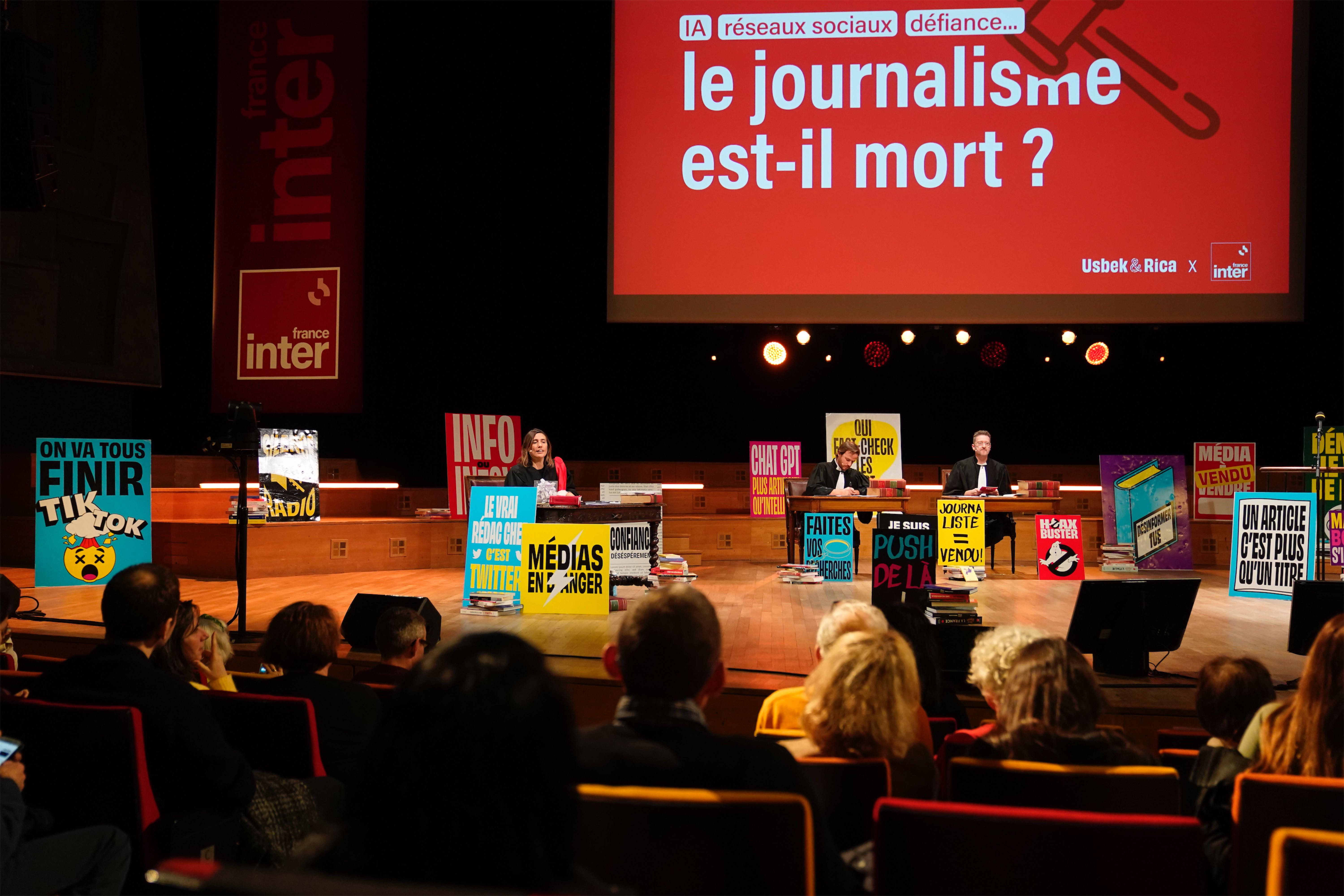 Vue d’ensemble d’une scène de débat dans un auditorium. Trois bureaux, des intervenants, des affiches sur les médias, les réseaux sociaux et la désinformation. En fond, l’écran affiche « Le journalisme est-il mort ? ».