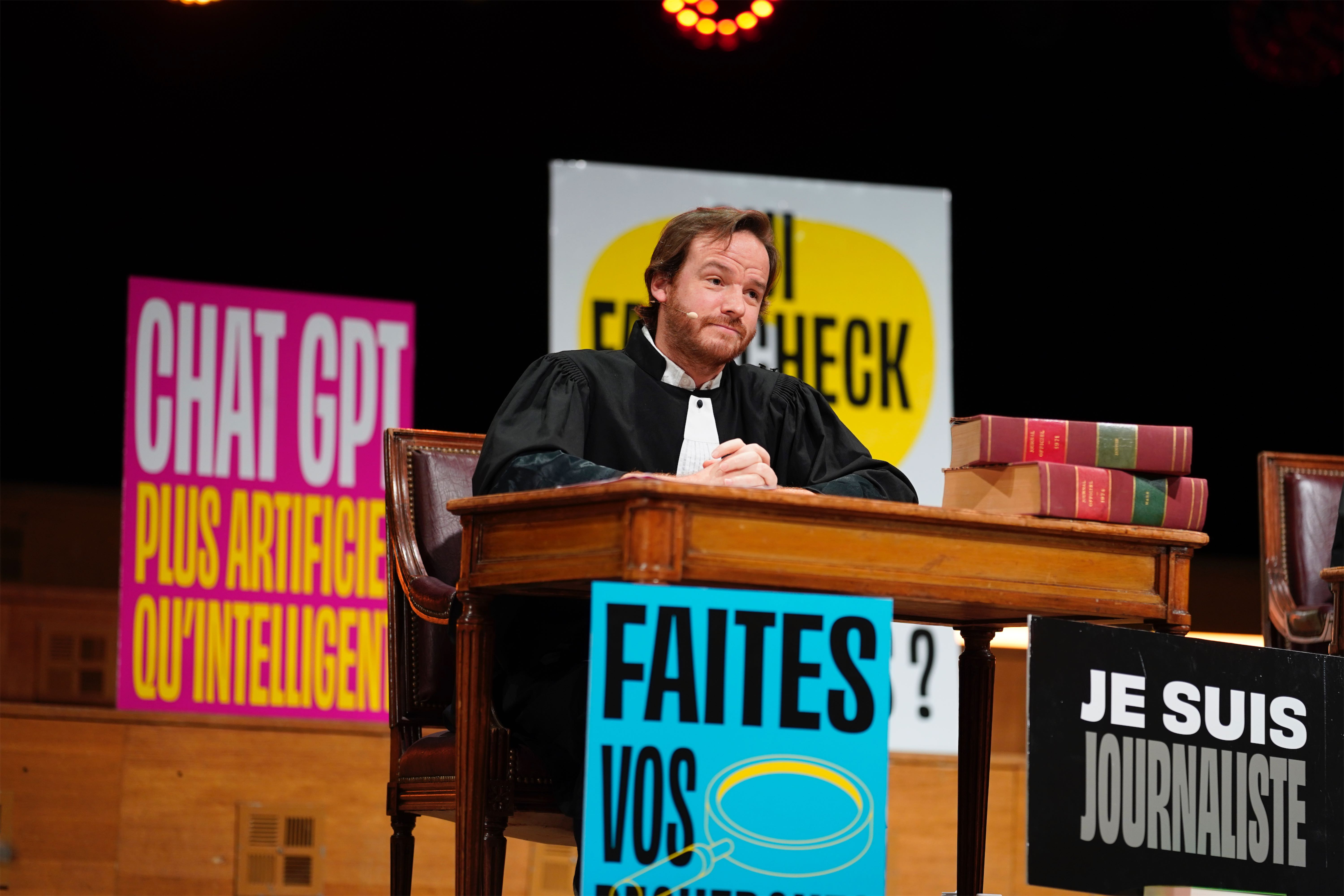 Gros plan d’un homme déguisé en juge, assis à un bureau, entouré de pancartes colorées dont l’une dit « Faites vos recherches » et une autre « Chat GPT plus artificiel qu’intelligent ».