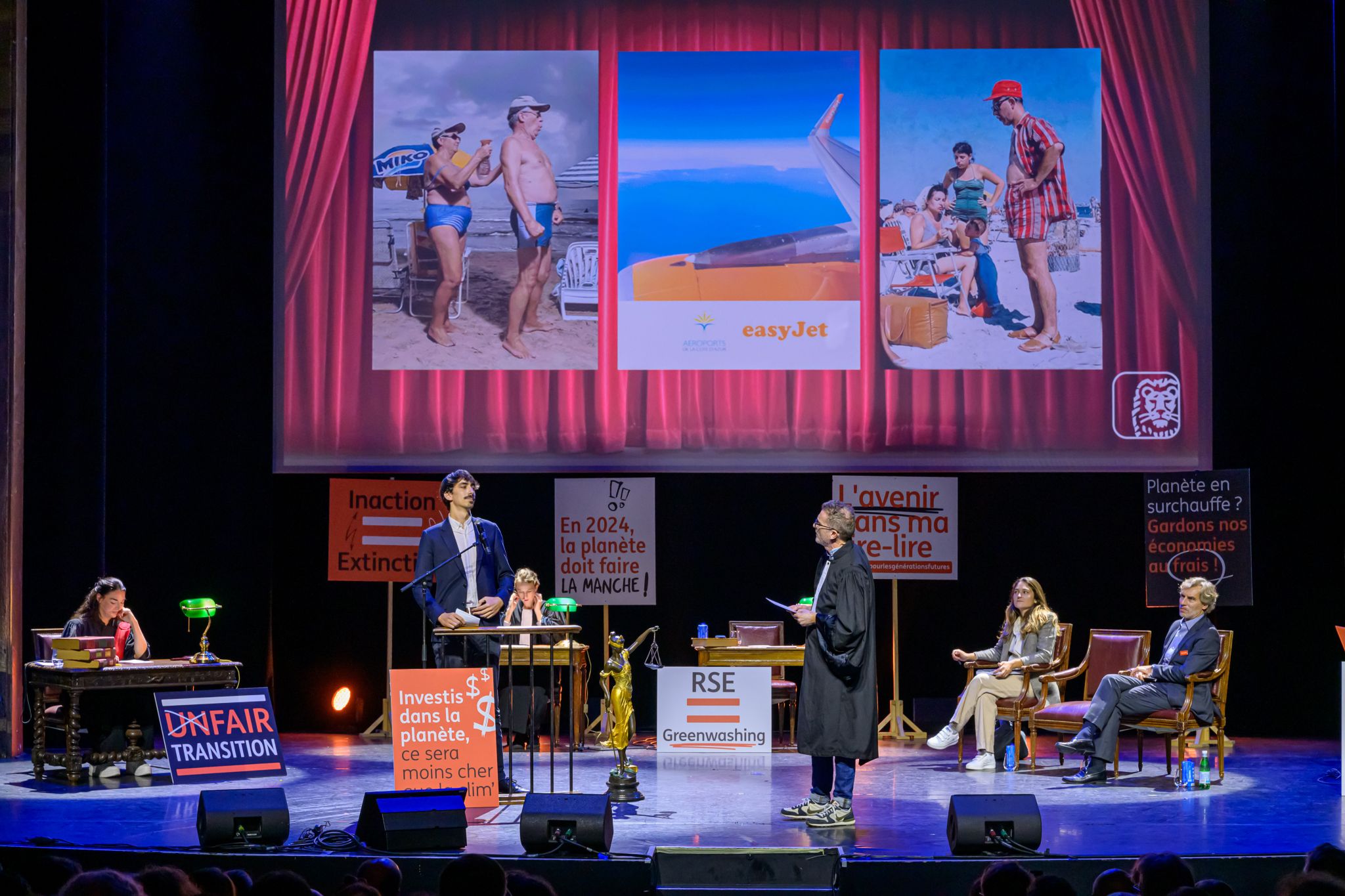 Mise en scène d’un procès fictif sur scène avec plusieurs intervenants. Trois grandes images de touristes à la plage et d’un avion sont projetées en fond. Des pancartes évoquant le climat et la transition sont visibles sur scène.