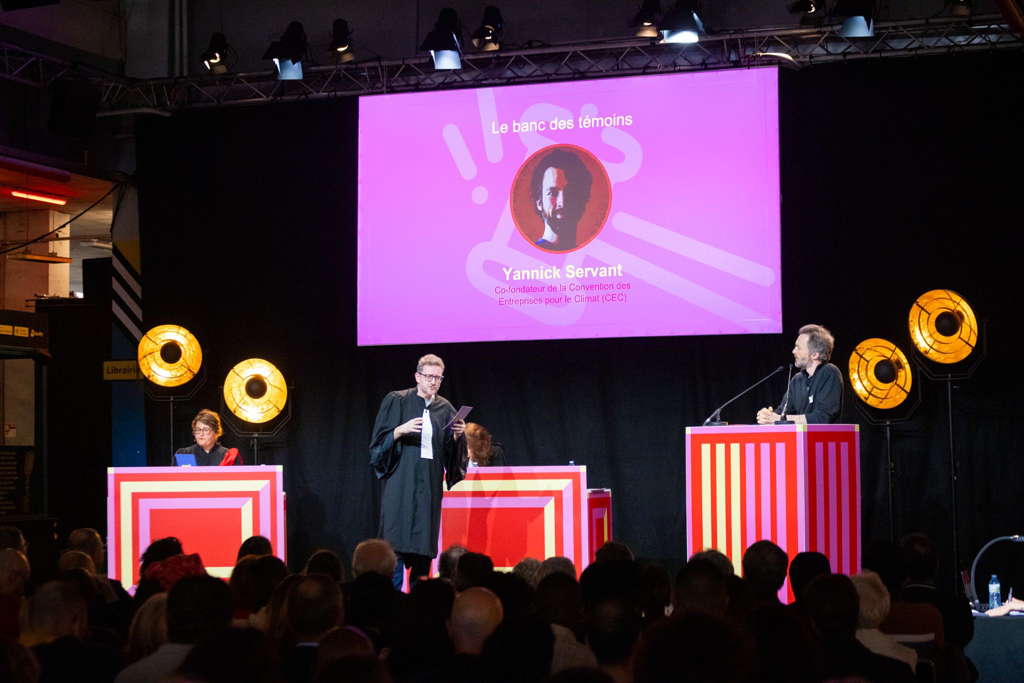 Scène sur une scène de débat avec trois pupitres rouges. Une personne habillée en avocat s’adresse à un homme debout derrière un pupitre, tandis qu’une troisième personne est assise à gauche. Un écran affiche le portrait de Yannick Servant avec la mention "Le banc des témoins".