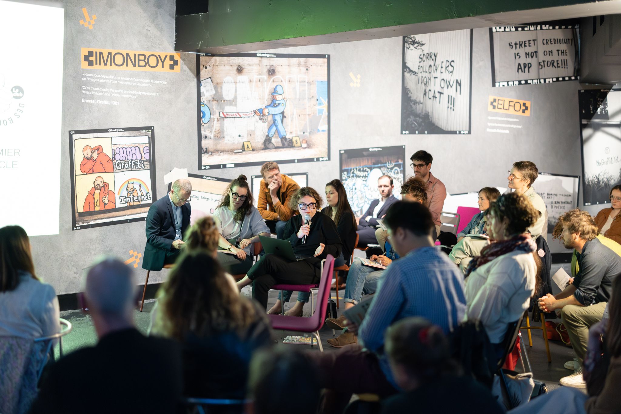 Vue d’ensemble d’un débat participatif, où des intervenants et membres du public échangent, entourés d’affiches de street art et de graffitis dans un espace culturel.