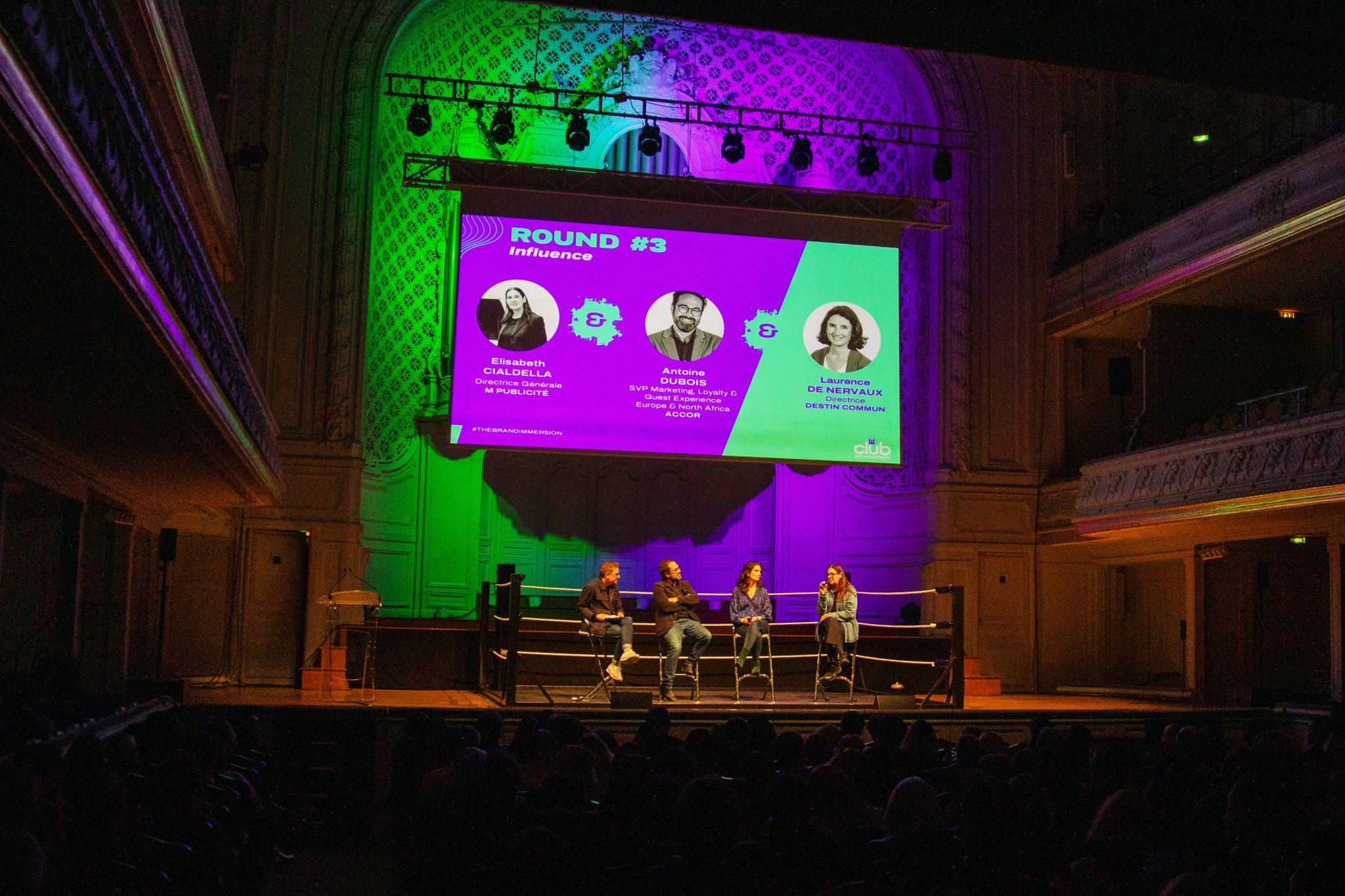 Une scène de théâtre éclairée en vert et violet accueille quatre intervenants assis sur des tabourets devant un grand écran. Le visuel projeté indique un débat sur l’influence avec les portraits et noms des intervenants.