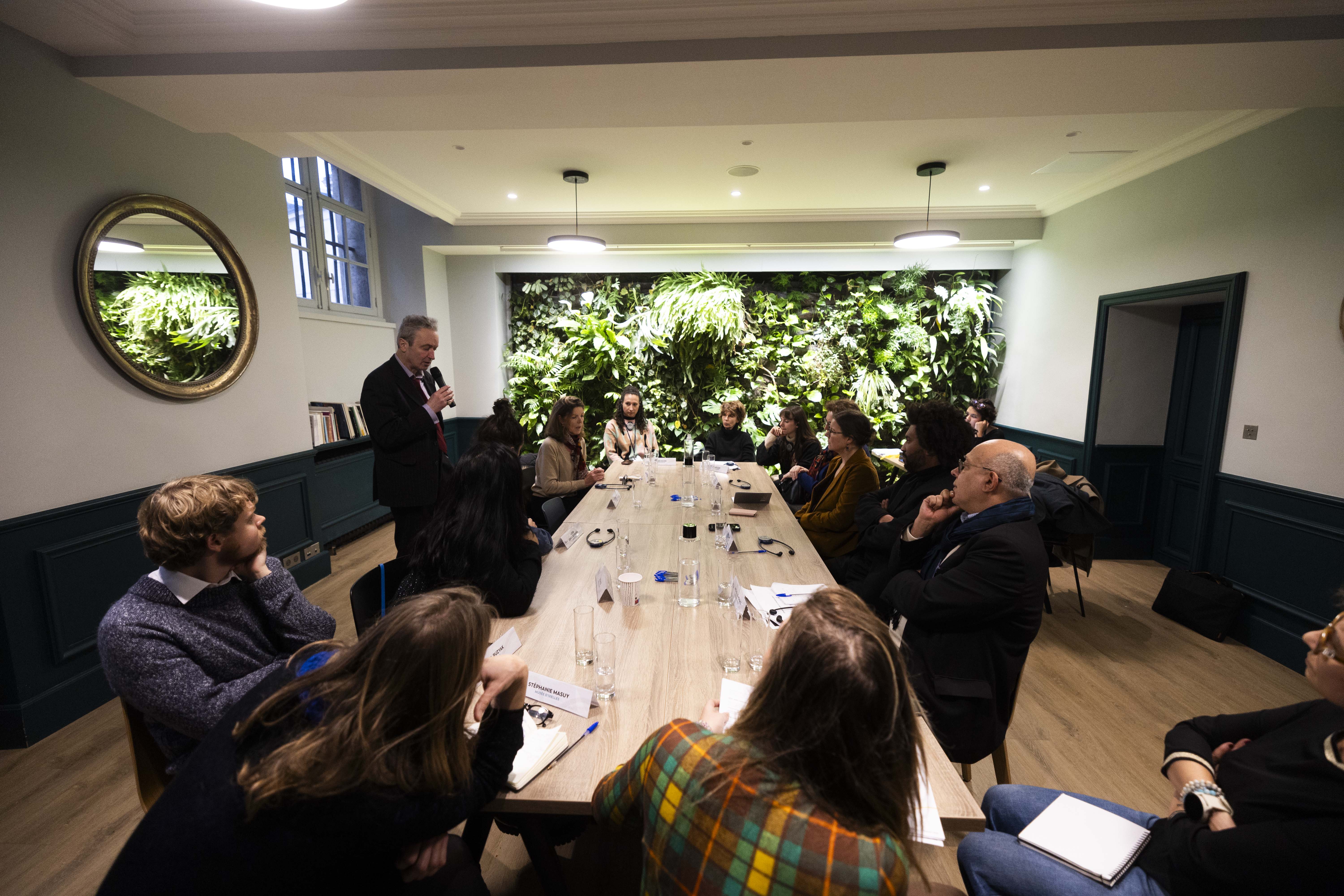 Une réunion en petit comité autour d’une grande table en bois, dans une salle élégante avec un mur végétal. Une personne debout prend la parole, pendant que les autres participants, assis, prennent des notes ou écoutent.