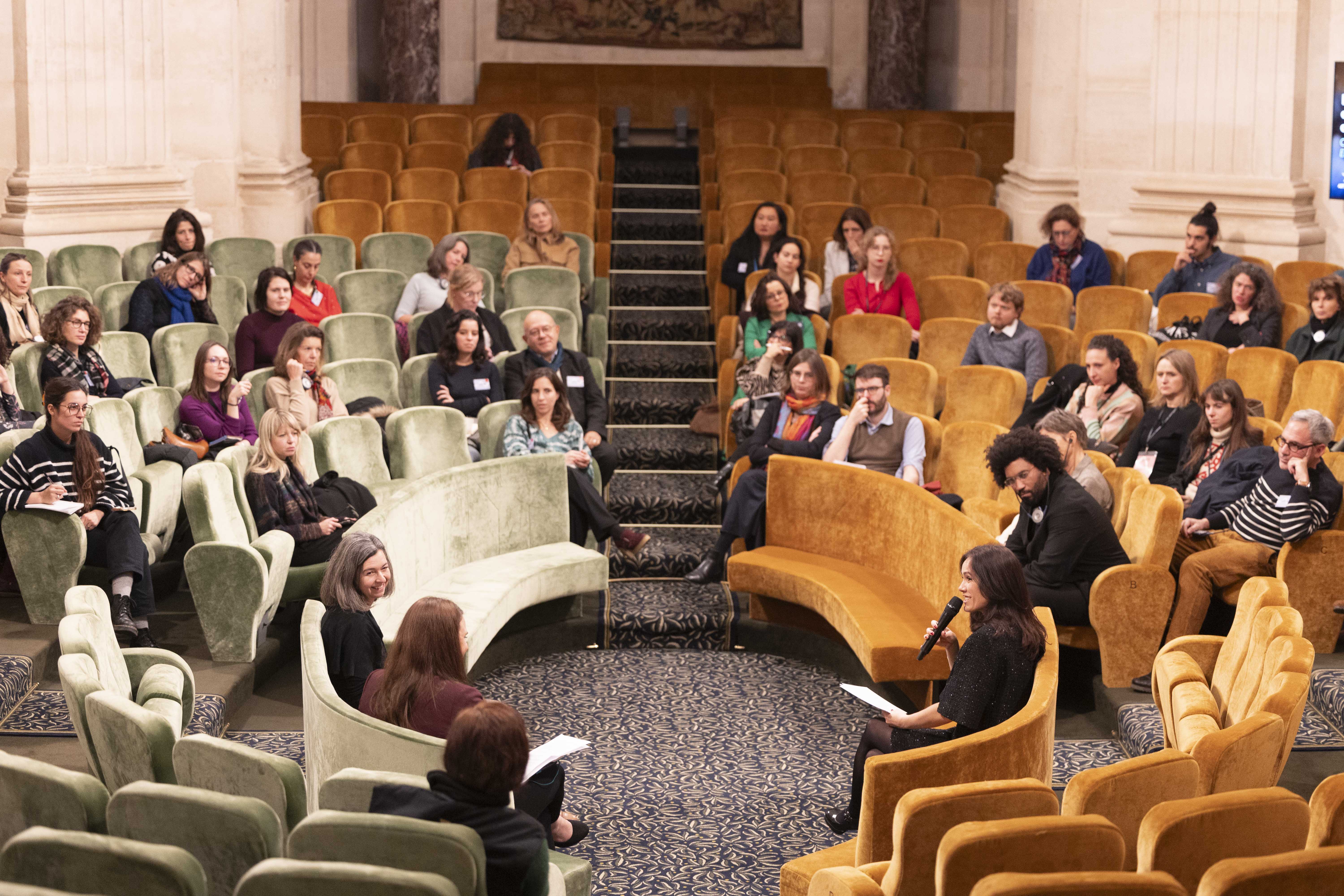Une conférence se déroule dans un amphithéâtre aux sièges colorés, verts et jaunes. Une femme au premier plan s’adresse au public avec un micro, tandis que l’audience, répartie sur plusieurs rangées, l’écoute attentivement.