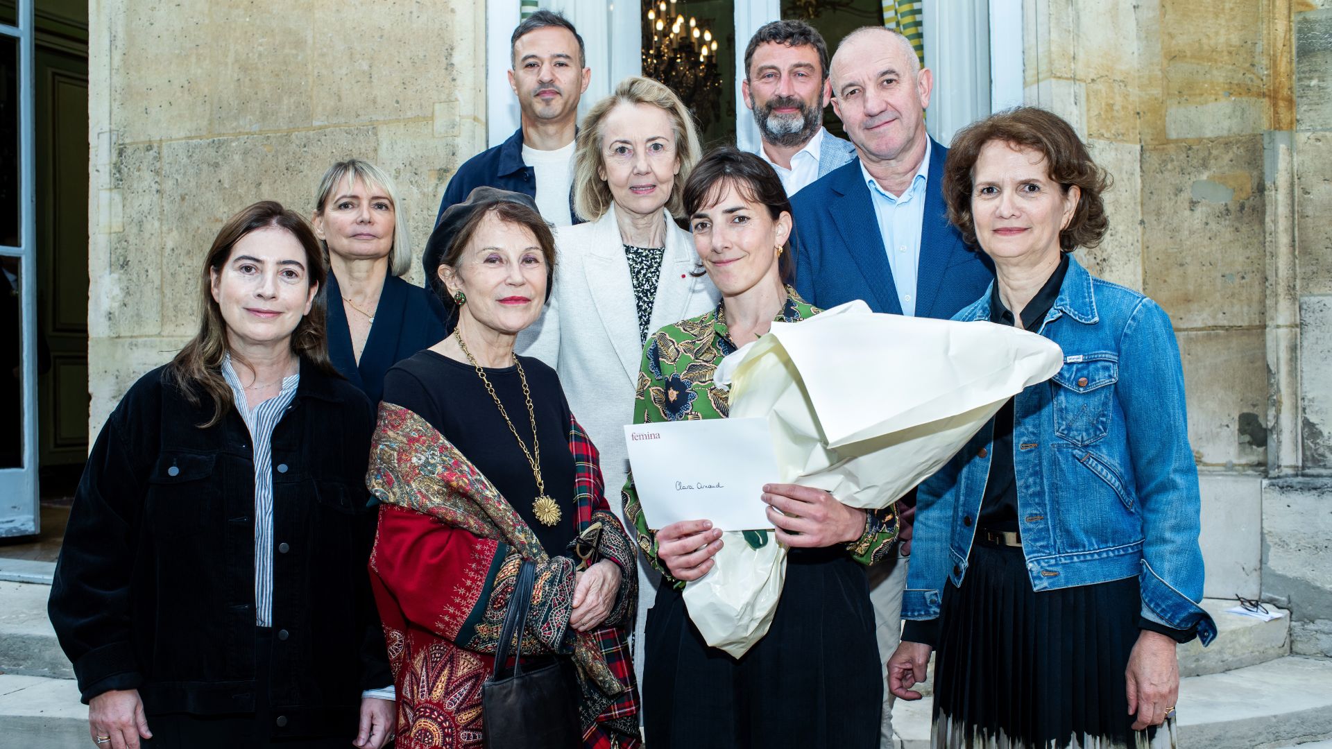 Un groupe de dix personnes, hommes et femmes, pose ensemble à l’extérieur devant une façade en pierre claire. Une femme au centre tient un grand bouquet de fleurs et une enveloppe. L’ambiance est formelle et bienveillante.