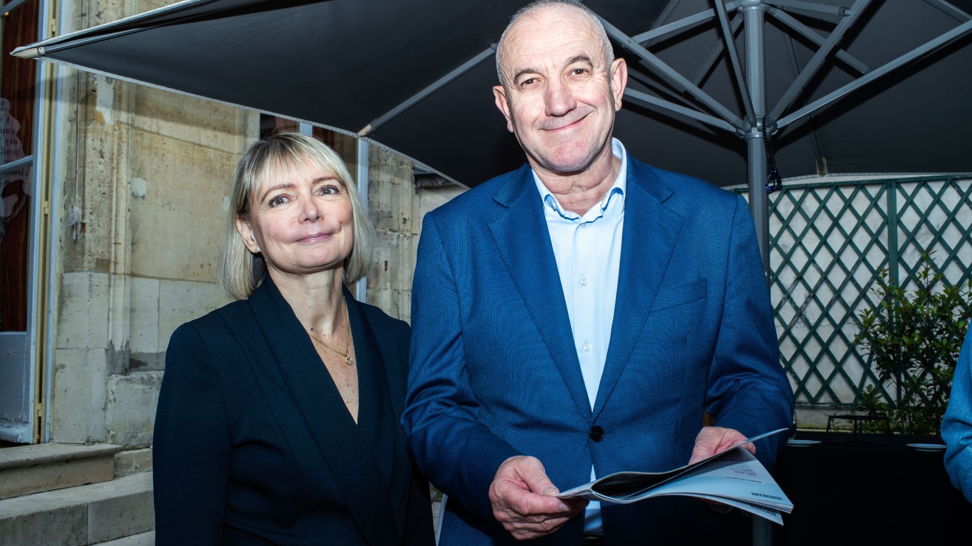 Anne Michelet et un homme posent côte à côte en souriant sous un grand parasol, devant un bâtiment en pierre. L’homme tient un livret ouvert dans les mains.