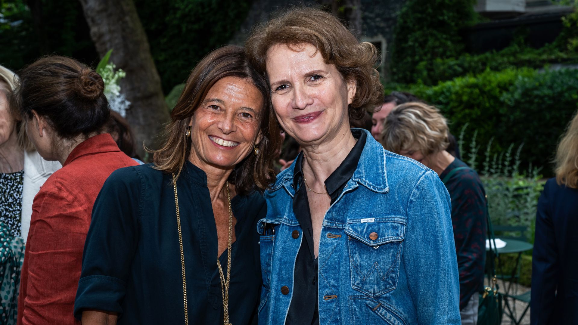 Deux femmes souriantes posent tête contre tête dans un jardin. L’une porte une chemise bleu marine, l’autre une veste en jean. L’arrière-plan montre d'autres personnes en conversation.