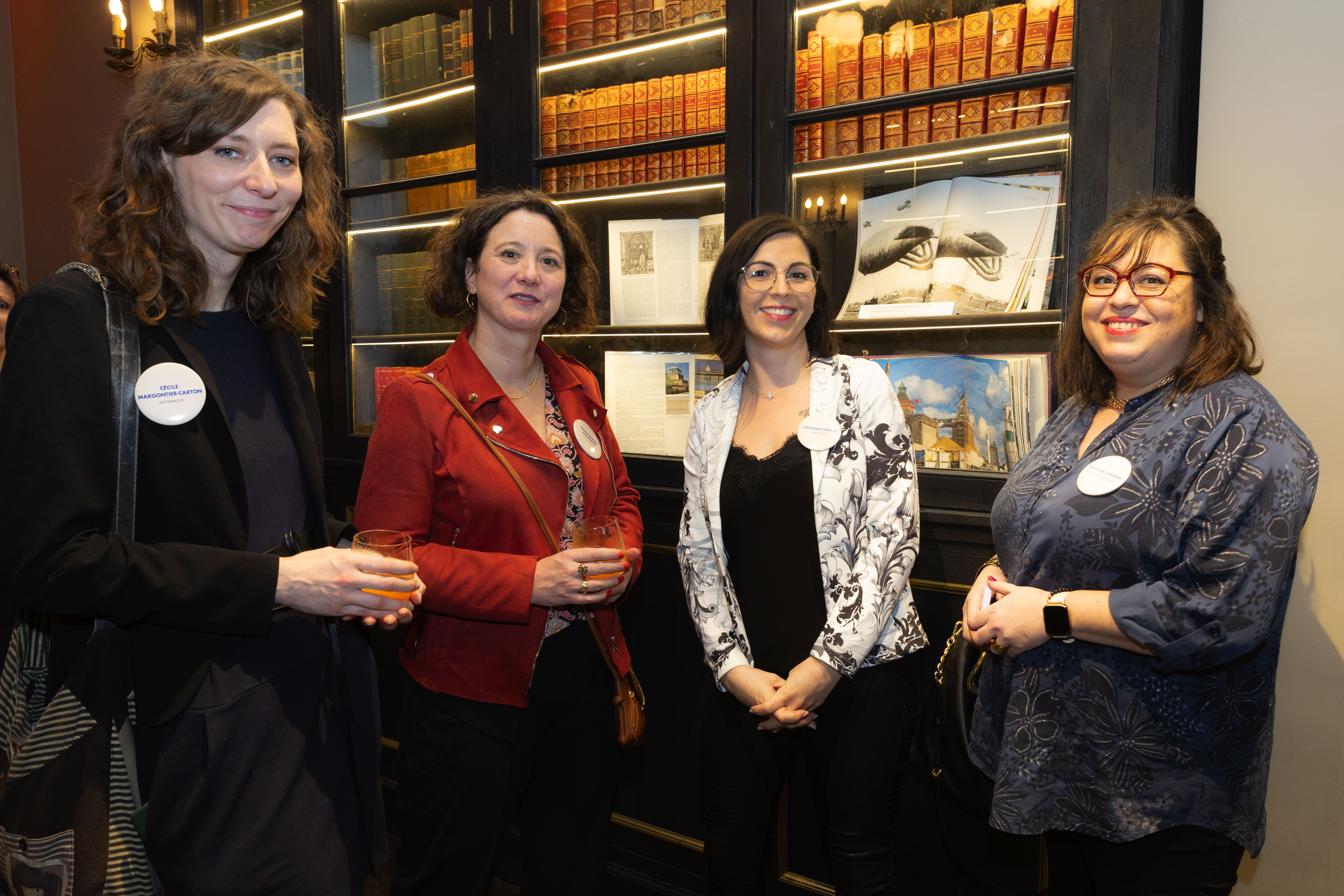 Quatre femmes posent debout, un verre à la main, devant une bibliothèque contenant des livres anciens. Elles portent des badges d’identification, suggérant leur participation à un événement professionnel ou associatif.