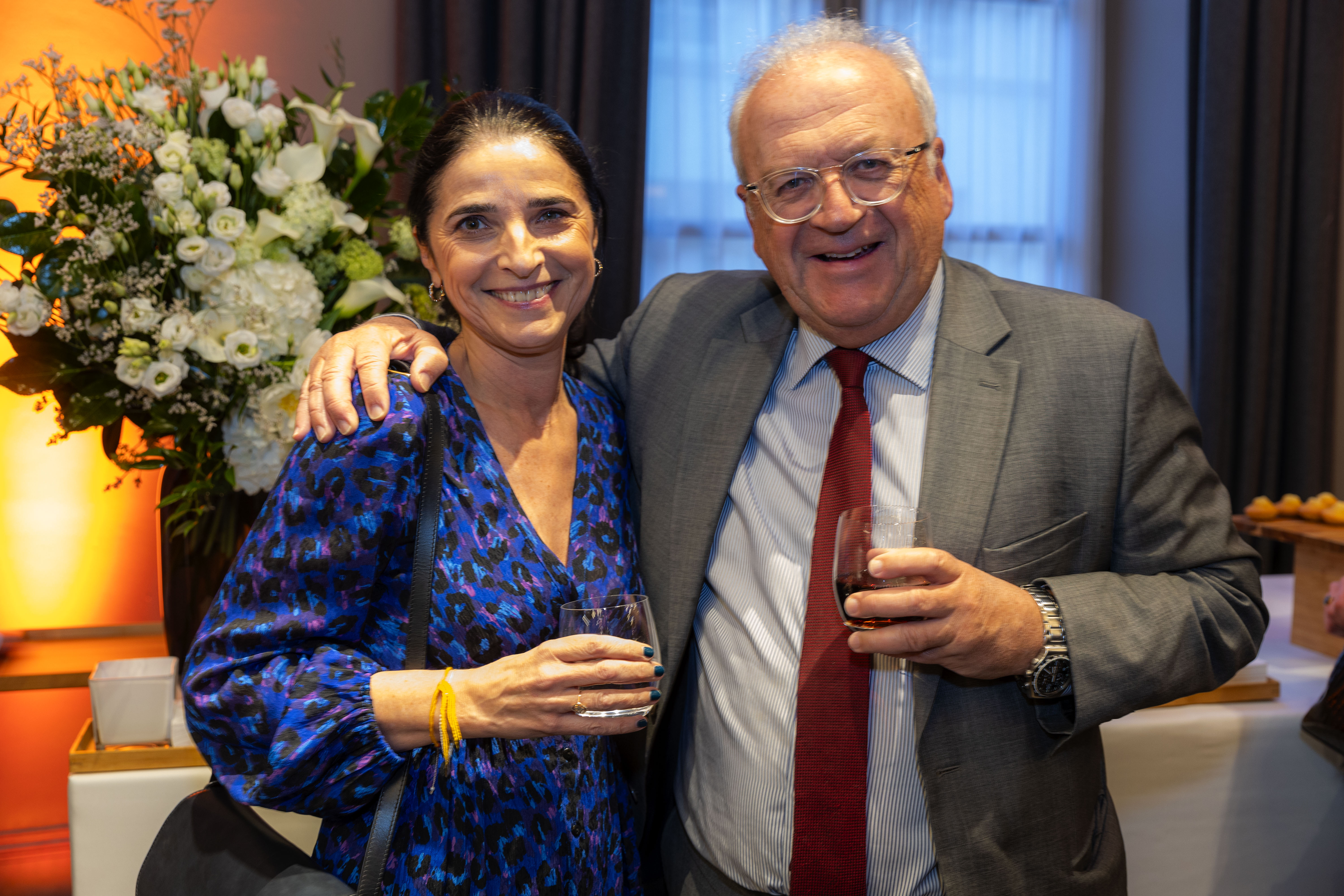 Valérie Salomon et un homme, souriants, posent ensemble en tenue élégante avec un verre à la main, devant une composition florale blanche dans un cadre formel, probablement un cocktail ou une remise de prix.
