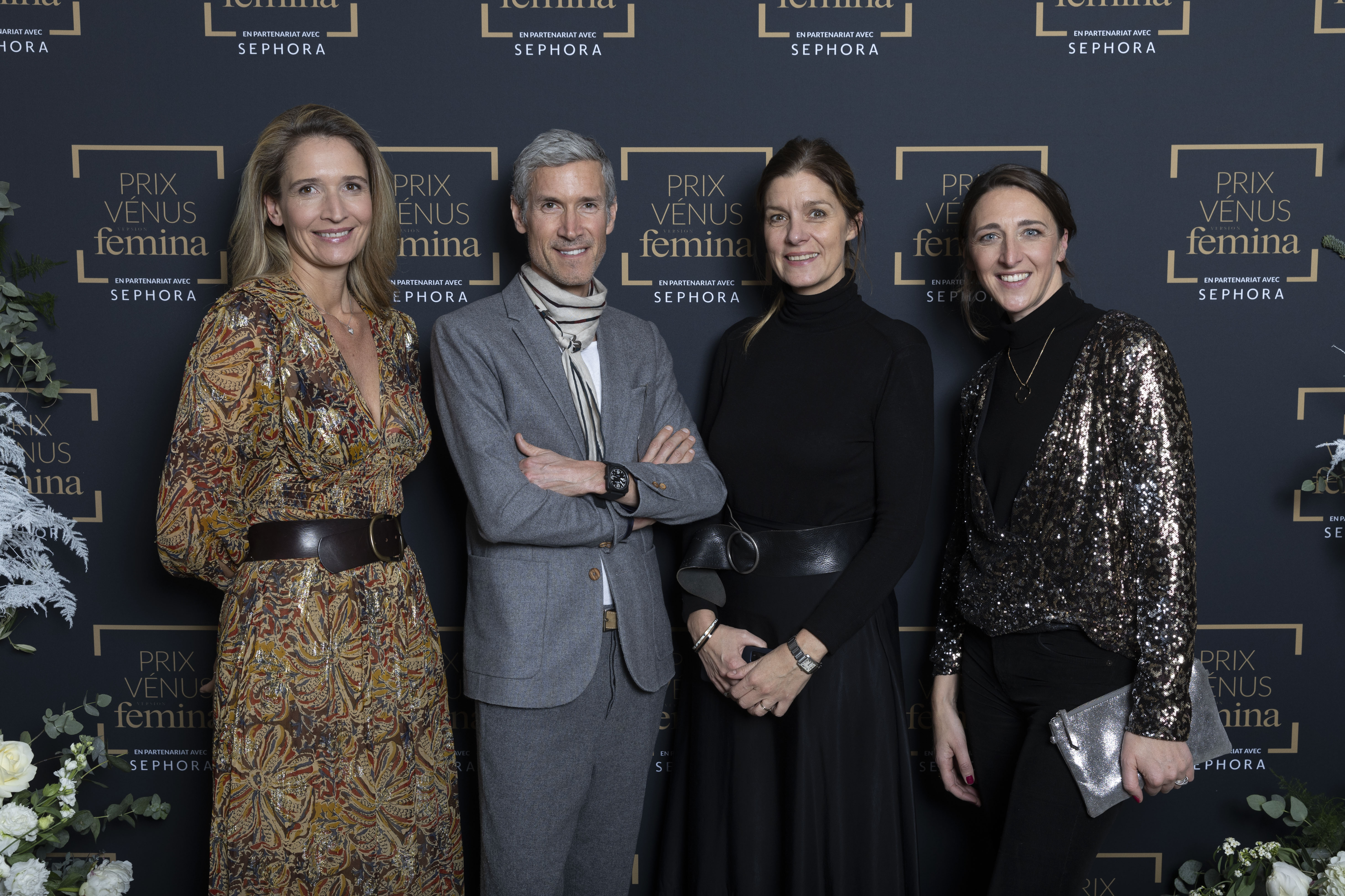 Quatre personnes posent devant le mur du Prix Vénus Femina. Trois femmes et un homme en tenue chic et élégante, souriants, entourés de décorations florales.