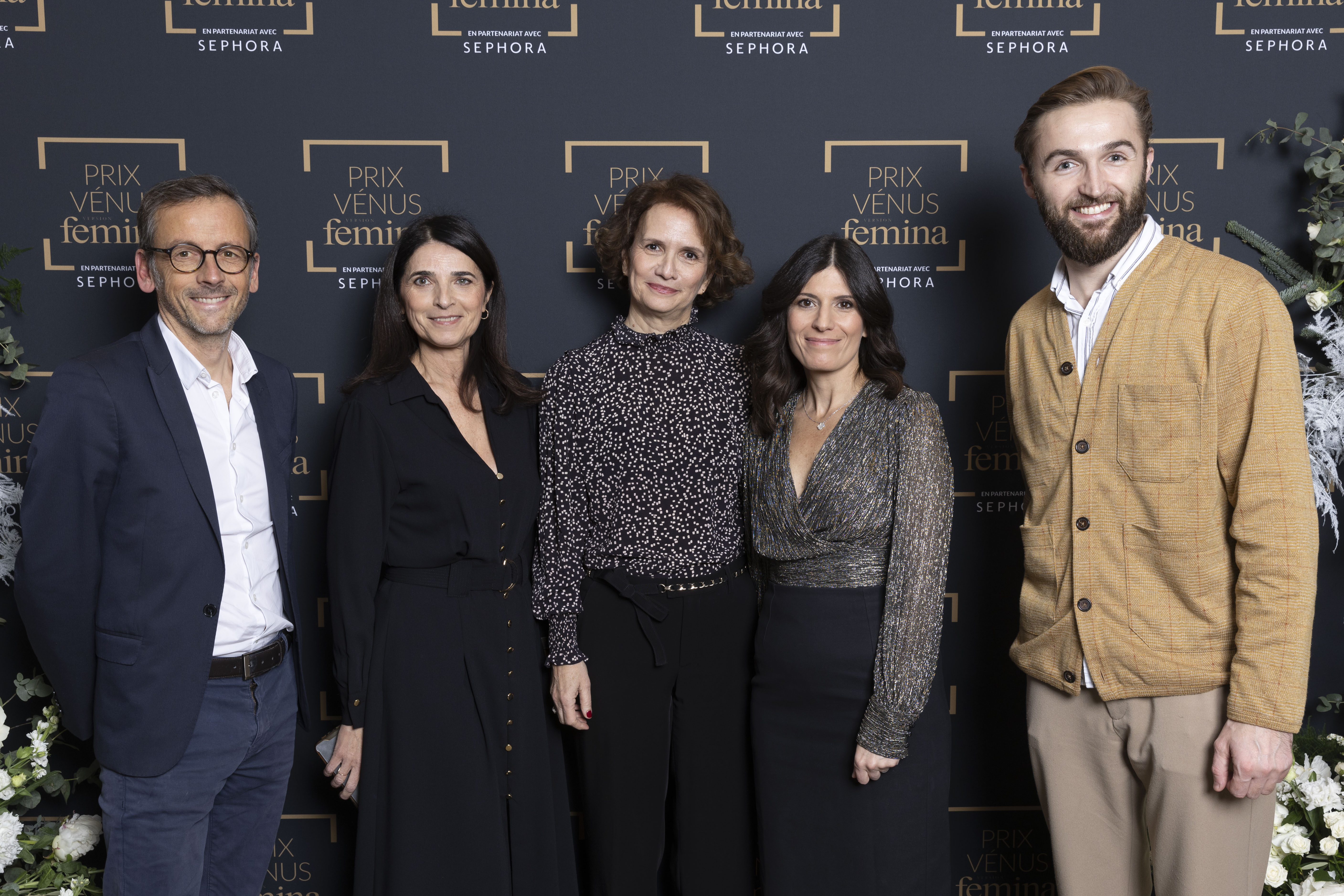 Groupe de cinq personnes élégamment habillées posant devant le mur officiel du Prix Vénus Femina. On retrouve trois femmes et deux hommes, tous souriants.