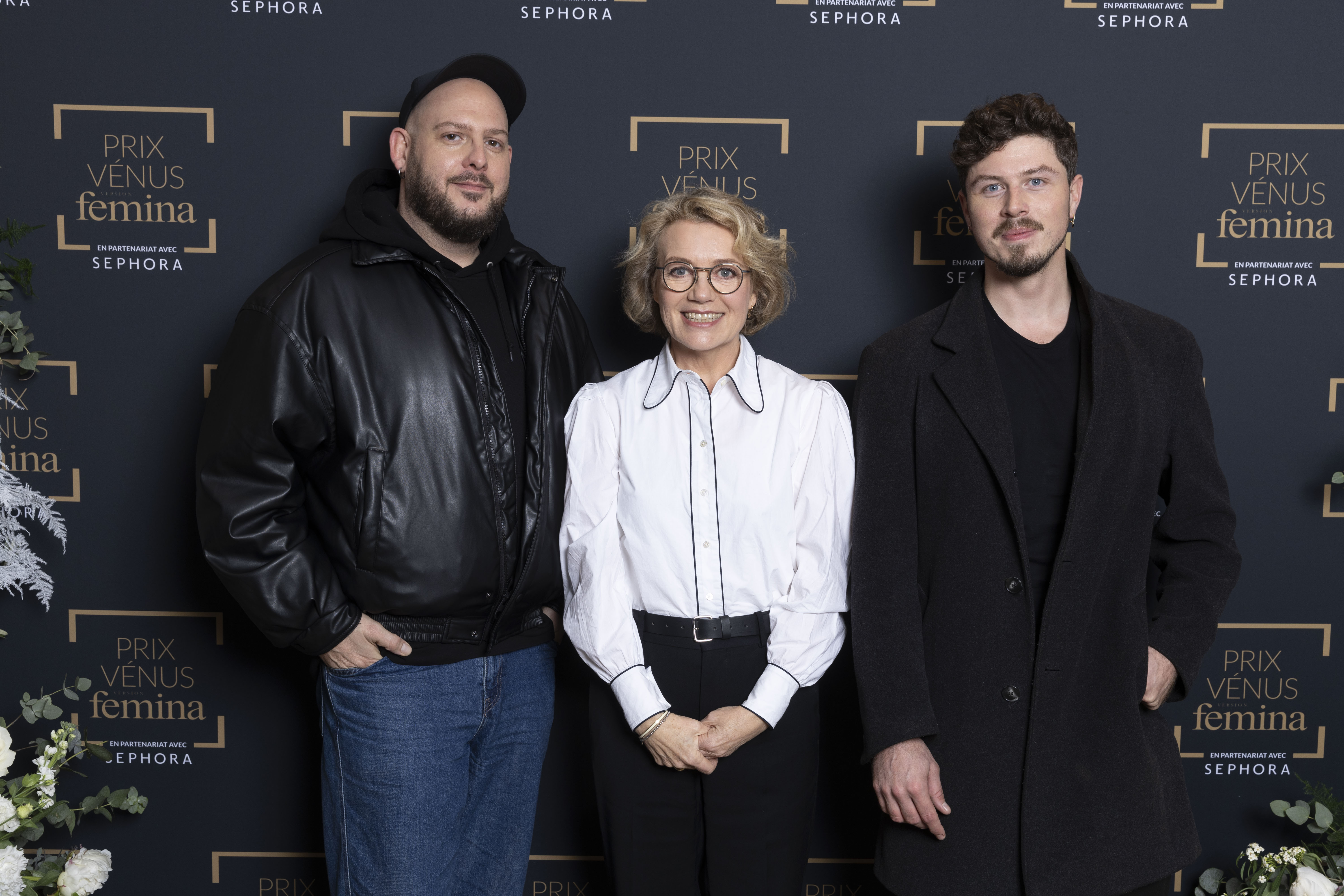 Trois personnes posent devant un fond noir marqué du logo du Prix Vénus Femina. La personne au centre porte une chemise blanche et les deux hommes de chaque côté sont habillés en noir, l’un avec une veste en cuir, l’autre en manteau long