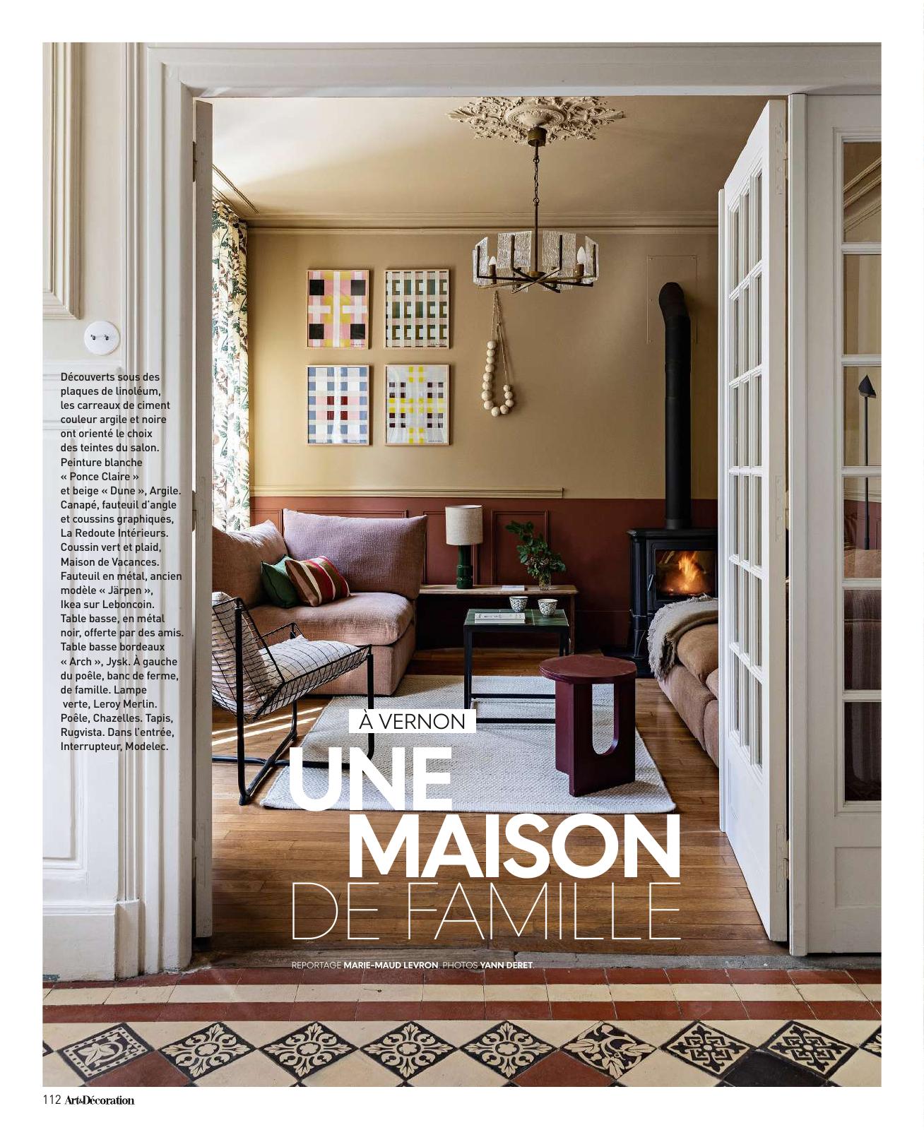 Page intérieure du magazine Art & Décoration, présentant un salon chaleureux dans une maison familiale à Vernon. On y voit une pièce décorée dans des tons argile, beige et noir, avec un canapé d’angle brun, un fauteuil en métal noir et blanc, une table basse bordeaux, un tapis clair et un poêle noir à bois allumé. Des carreaux de ciment géométriques ornent l’entrée de la pièce. Sur les murs, quatre tableaux graphiques colorés sont accrochés au-dessus du canapé. Le titre en bas de page annonce : « À Vernon, une maison de famille ».