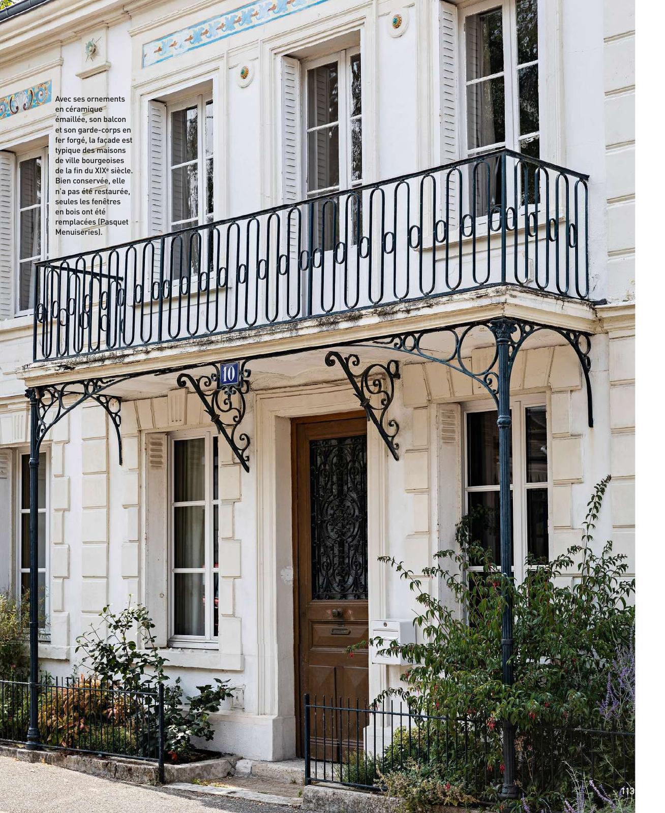 Façade d’une maison de ville bourgeoise du XIXe siècle, bien conservée, avec un balcon et une marquise en fer forgé aux motifs décoratifs. Les encadrements des fenêtres sont ornés de céramiques émaillées bleues et beiges. La porte d’entrée en bois est encadrée de fenêtres à petits carreaux et d’un garde-corps noir. Des plantes et buissons bordent la façade.