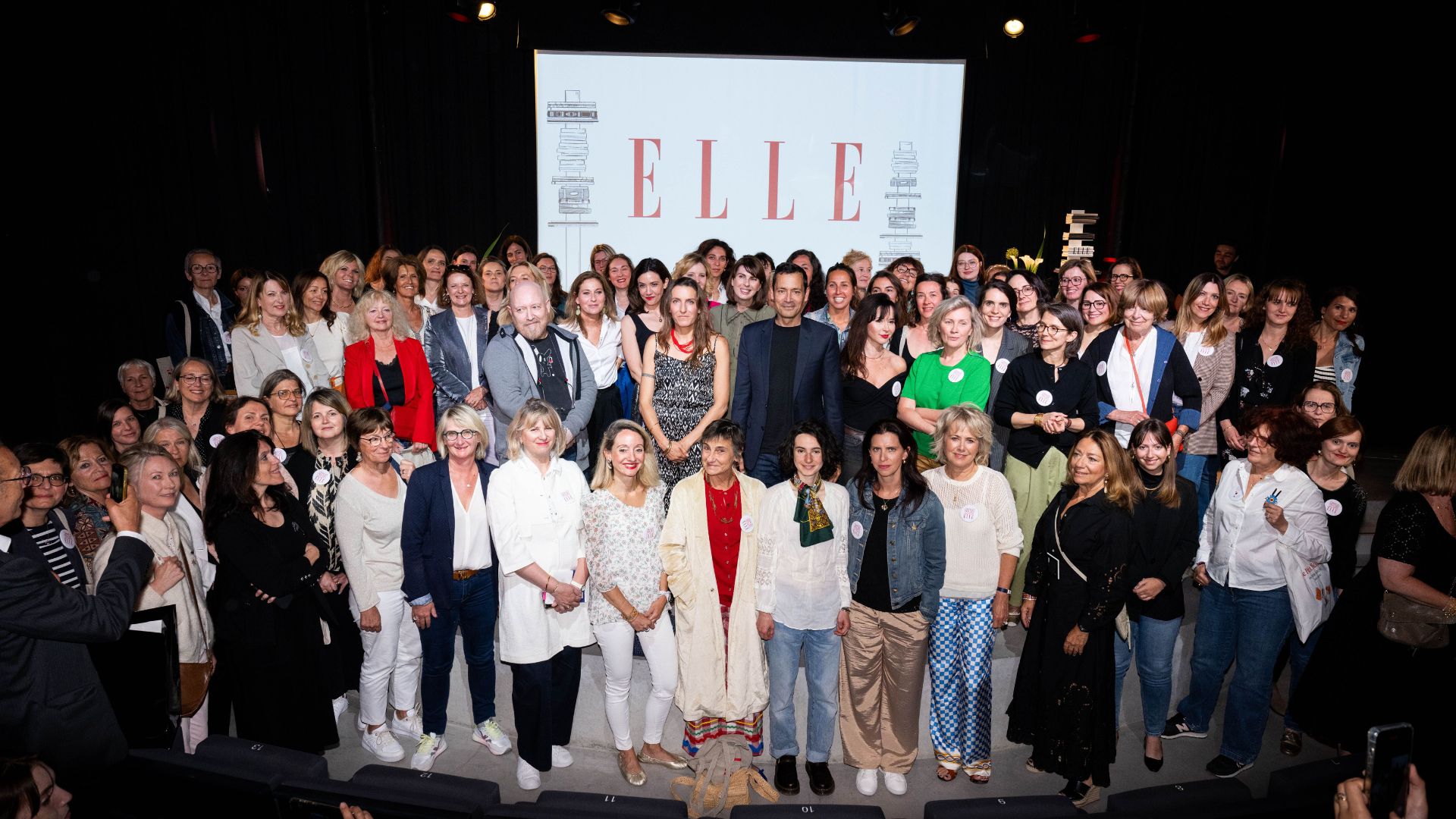 Un grand groupe de femmes et d'hommes est rassemblé sur scène devant un écran affichant le logo du magazine ELLE. Ils posent ensemble pour une photo de groupe, probablement à l’occasion d’un événement culturel ou littéraire. L’atmosphère est chaleureuse et collaborative.