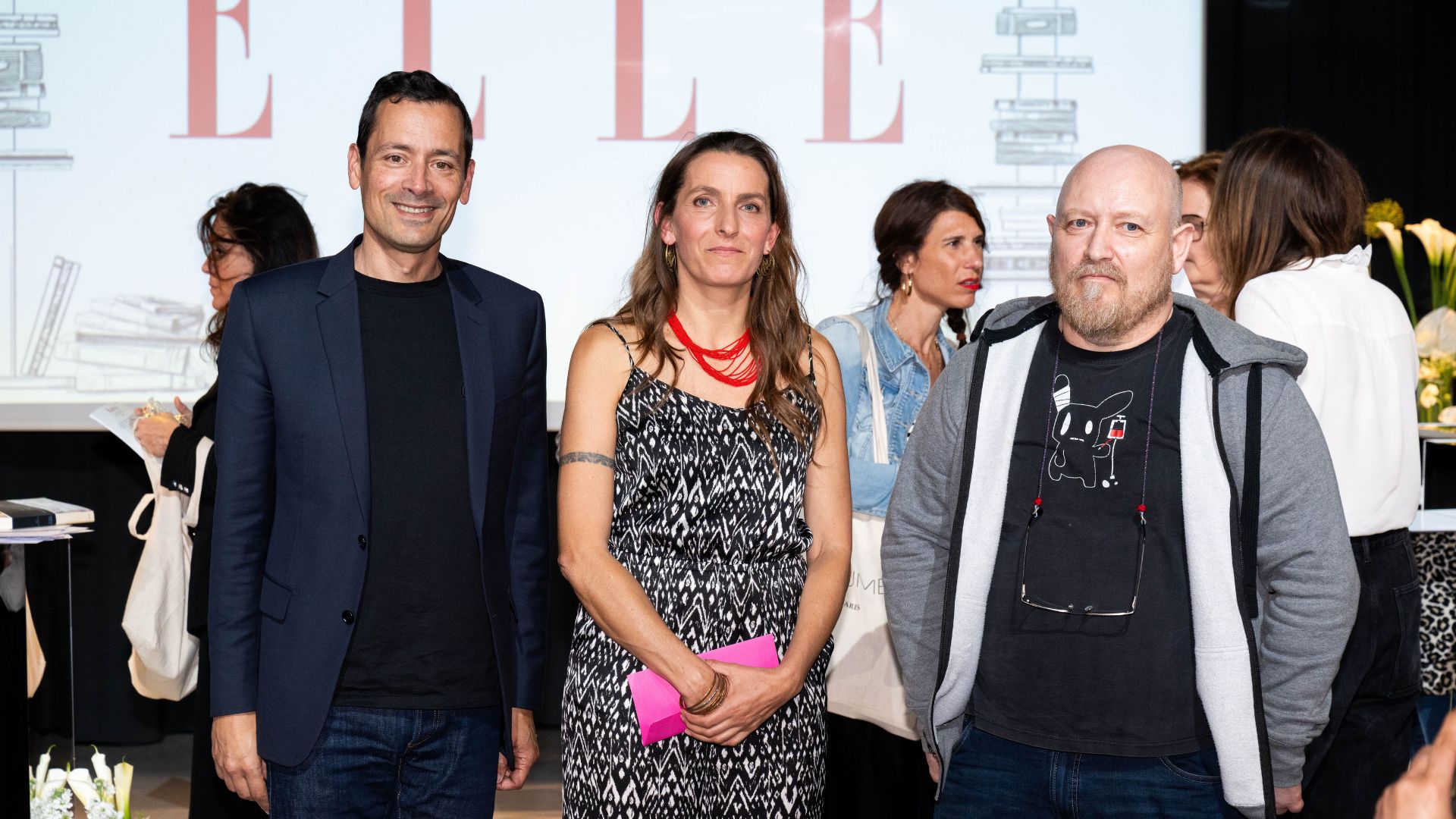 Trois personnes posent devant un écran arborant le logo ELLE : un homme en veste bleu marine, une femme portant une robe à motifs noirs et blancs avec un collier rouge, et un homme chauve avec une barbe, en t-shirt noir et sweat à capuche gris. L’événement semble être en lien avec le monde de la littérature ou de l’édition.