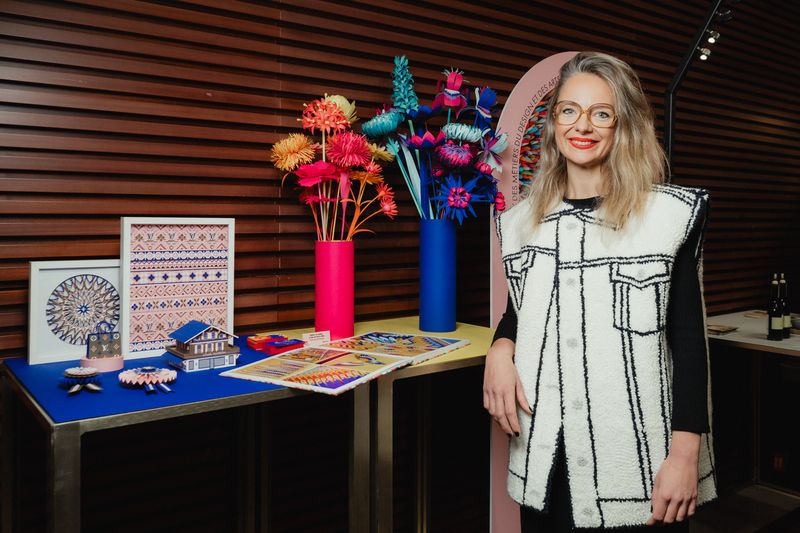 Portrait d’une femme blonde souriante portant des lunettes rondes et un gilet long blanc à imprimé noir, posant à côté d’un présentoir coloré. Sur la table : créations artisanales telles que fleurs en papier, maquette de maison, motifs brodés et objets décoratifs. En arrière-plan, deux grands vases vifs (rose et bleu) contiennent des fleurs artificielles multicolores. Cette scène se déroule dans le cadre du Prix des Artisanes, dédié à la valorisation du savoir-faire féminin.