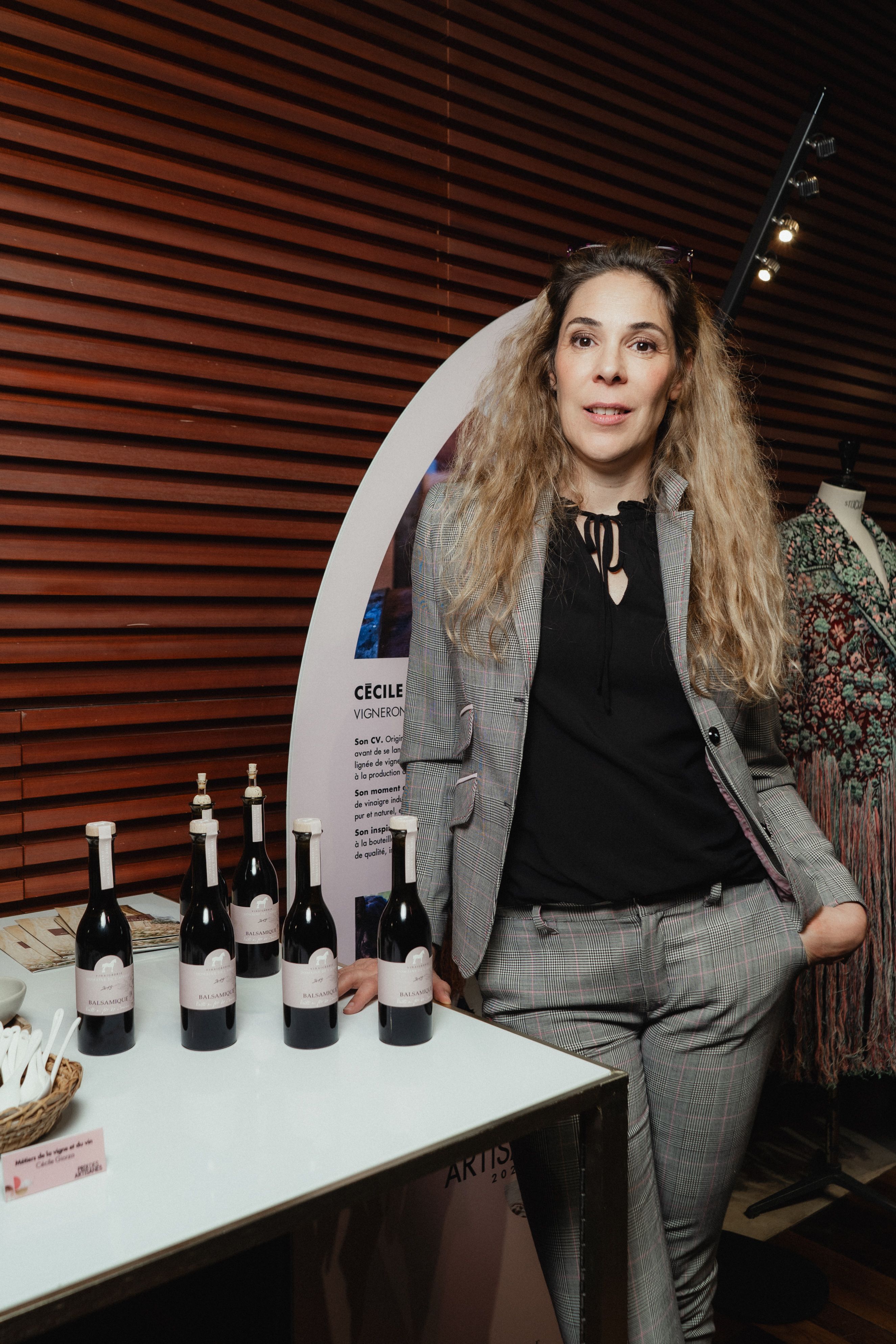 Portrait d’une femme aux cheveux longs et ondulés, habillée d’un costume à carreaux gris et noir. Elle se tient debout près d’une table exposant plusieurs bouteilles de vinaigre balsamique, toutes étiquetées « Balsamique ». En arrière-plan, un panneau présente son profil de vigneronne dans le cadre du Prix des Artisanes 2024. Un mannequin vêtu d’un vêtement brodé est visible à droite.