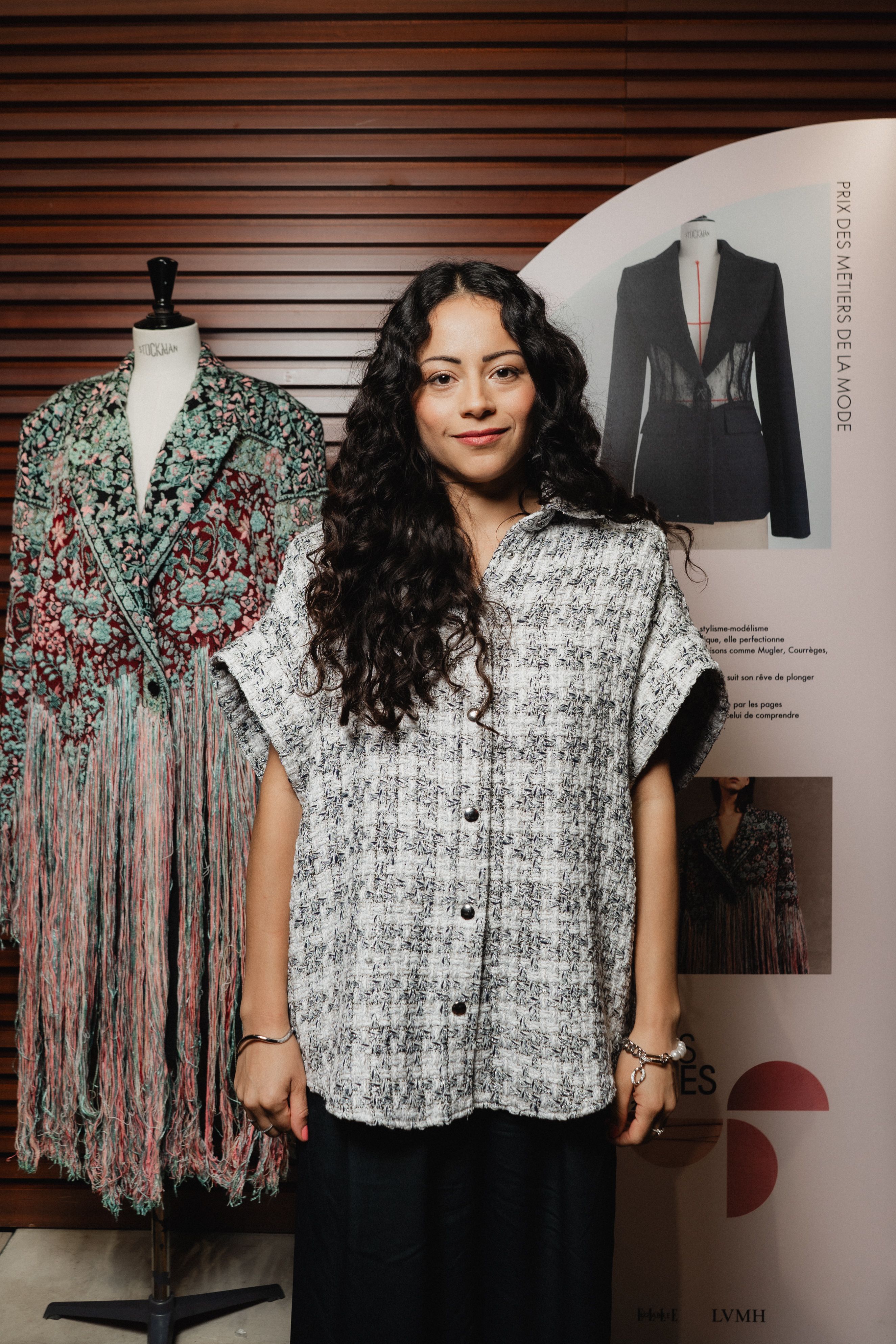 : Jeune femme aux cheveux longs et bouclés posant devant un panneau intitulé « Prix des Métiers de la Mode ». Elle porte une chemise à manches courtes en tissu texturé gris clair à boutons. Derrière elle, un mannequin expose un manteau brodé de fleurs rouges et vertes avec des franges multicolores. Le décor valorise le savoir-faire textile et la création artisanale féminine.