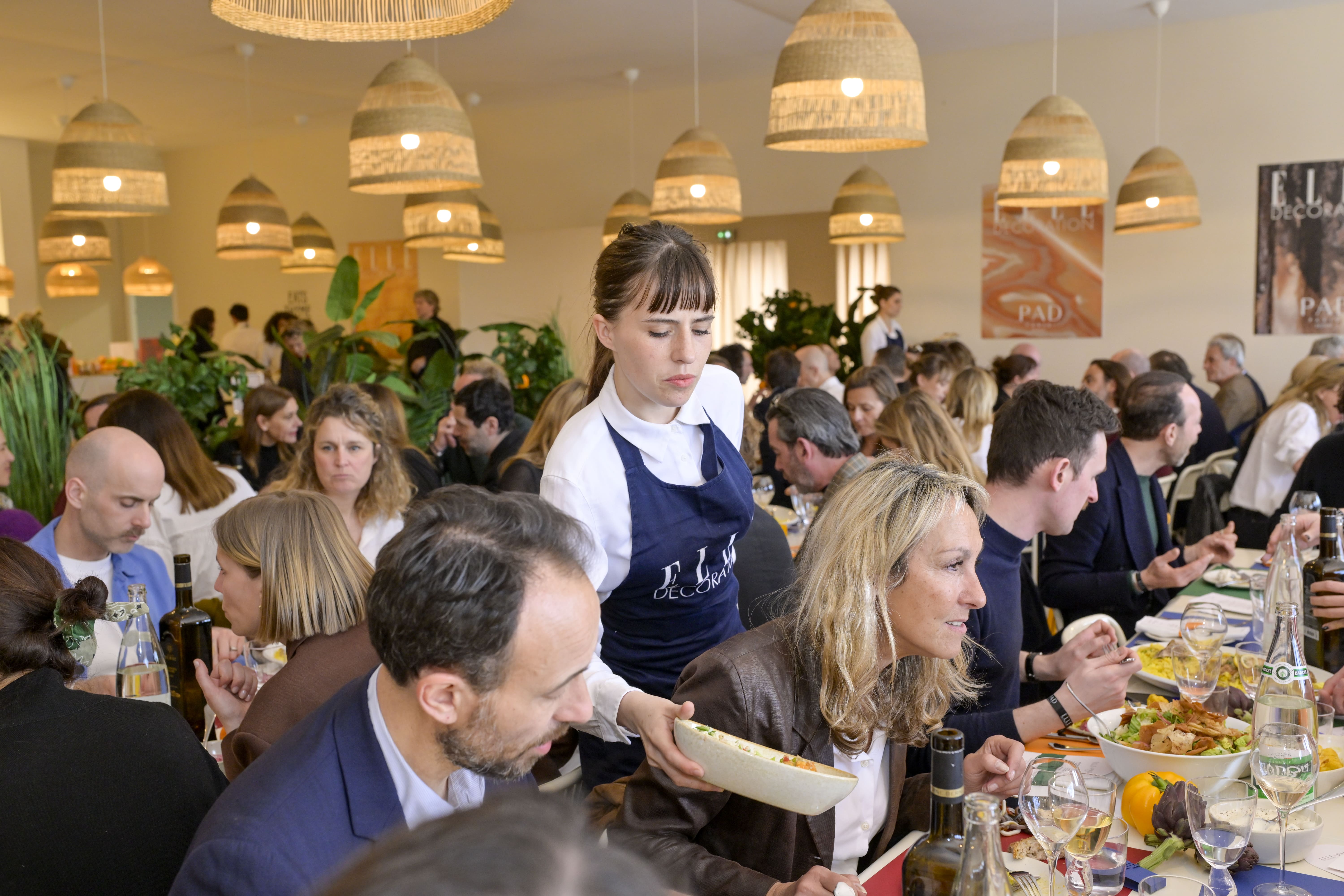 Une serveuse en tablier "ELLE Décoration" sert un plat à une grande tablée de convives. La salle, lumineuse et chaleureuse, est remplie d’invités assis à des tables décorées, entourées de plantes et de suspensions naturelles.