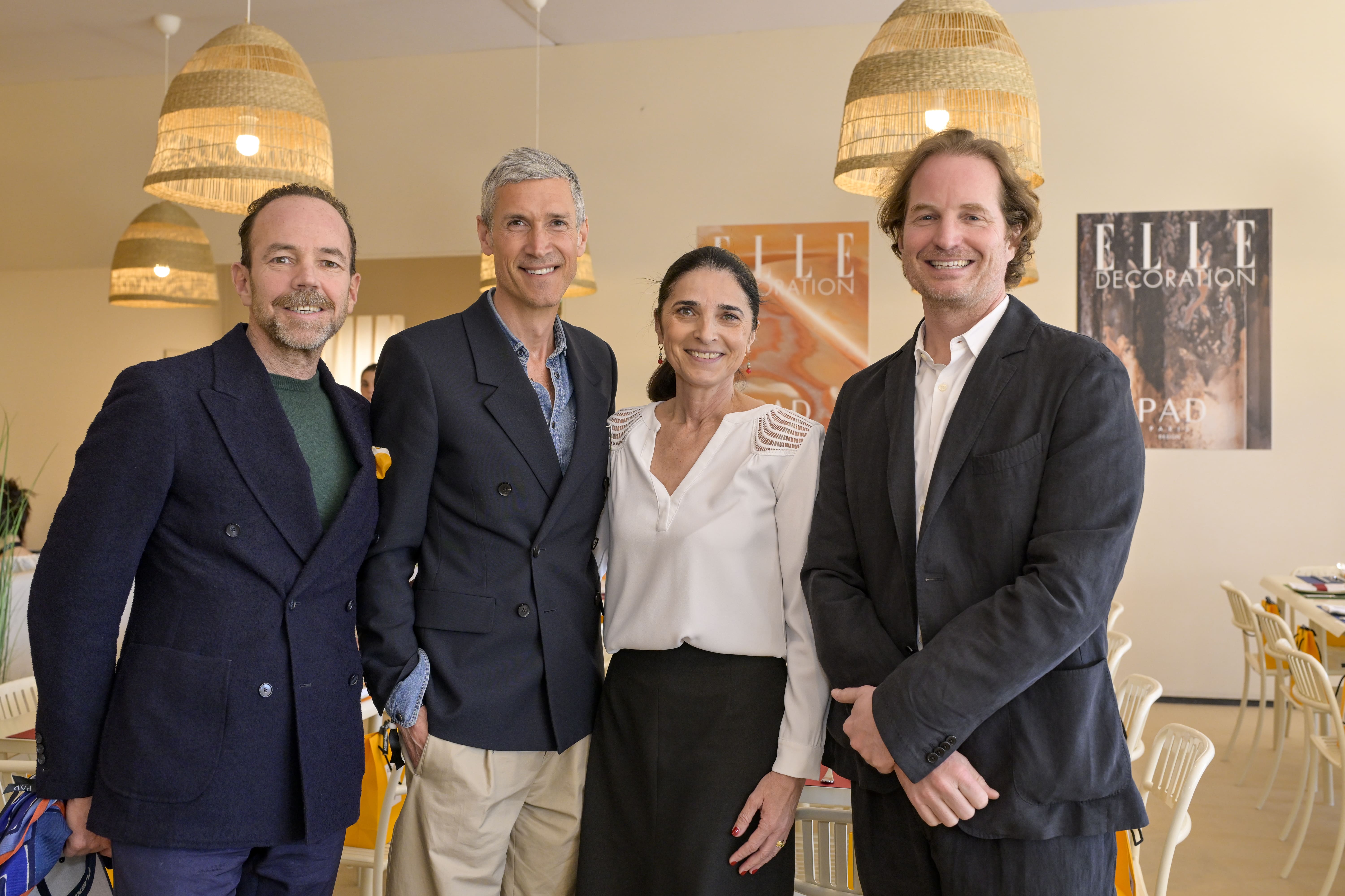 Trois hommes et une femme se tiennent debout, souriants, dans une salle décorée pour un déjeuner. La femme au centre porte une blouse blanche et les hommes sont en costume. En arrière-plan, les affiches ELLE Décoration sont accrochées aux murs couleur crème.