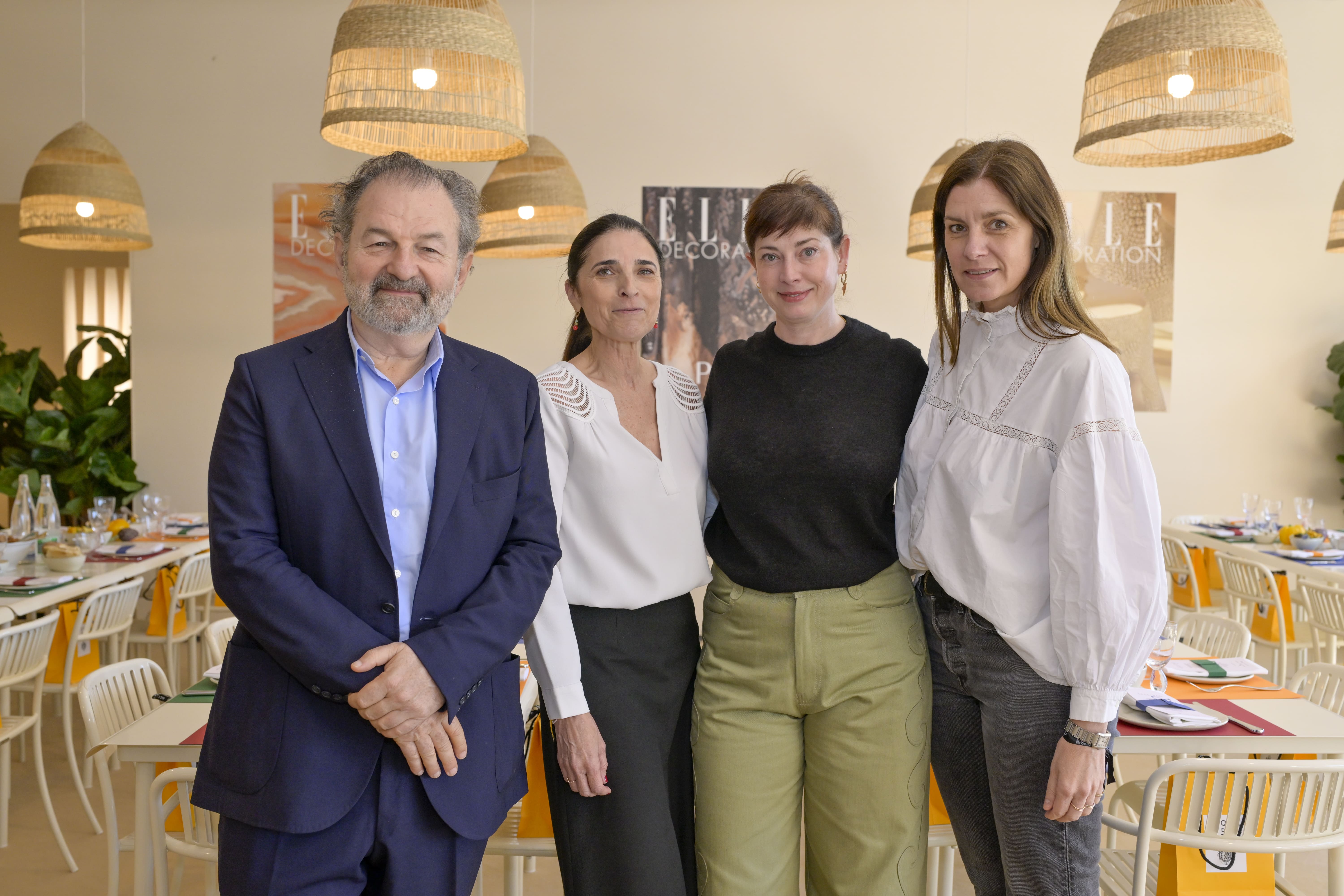 Quatre personnes posent ensemble dans une salle décorée de suspensions en rotin et de tables dressées pour un événement. En arrière-plan, des affiches ELLE Décoration sont visibles. De gauche à droite : un homme en costume bleu marine, une femme en blouse blanche et jupe noire, une femme en pull noir et pantalon vert clair, et une femme en chemise blanche à manches bouffantes.