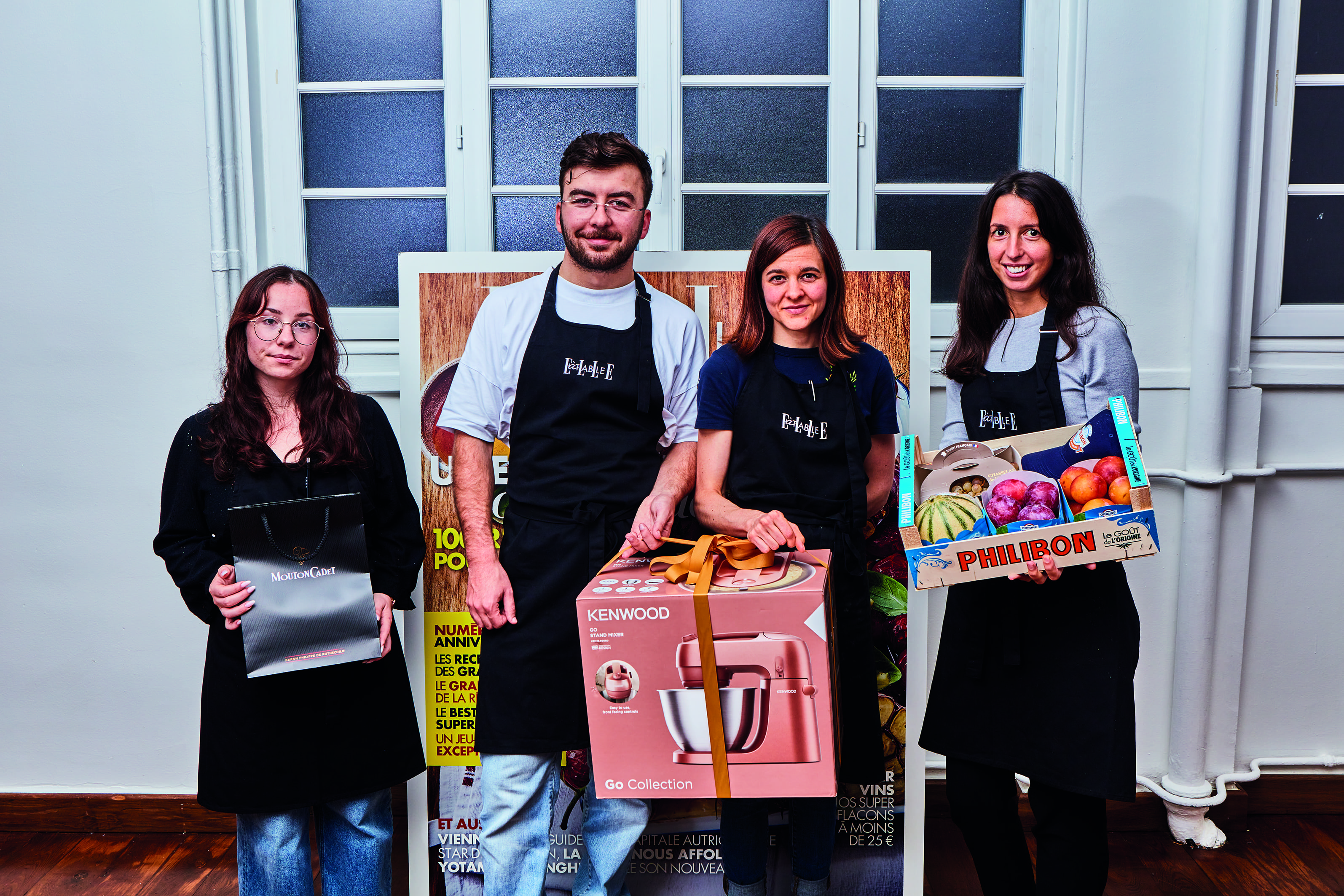 Quatre gagnant·es du concours E.A.T. posent debout devant une affiche ELLE à Table. Chacun·e tient un lot : un sac cadeau, un robot pâtissier Kenwood ou un panier de fruits de la marque Philibon. Ils portent tous un tablier noir floqué du logo du concours.
