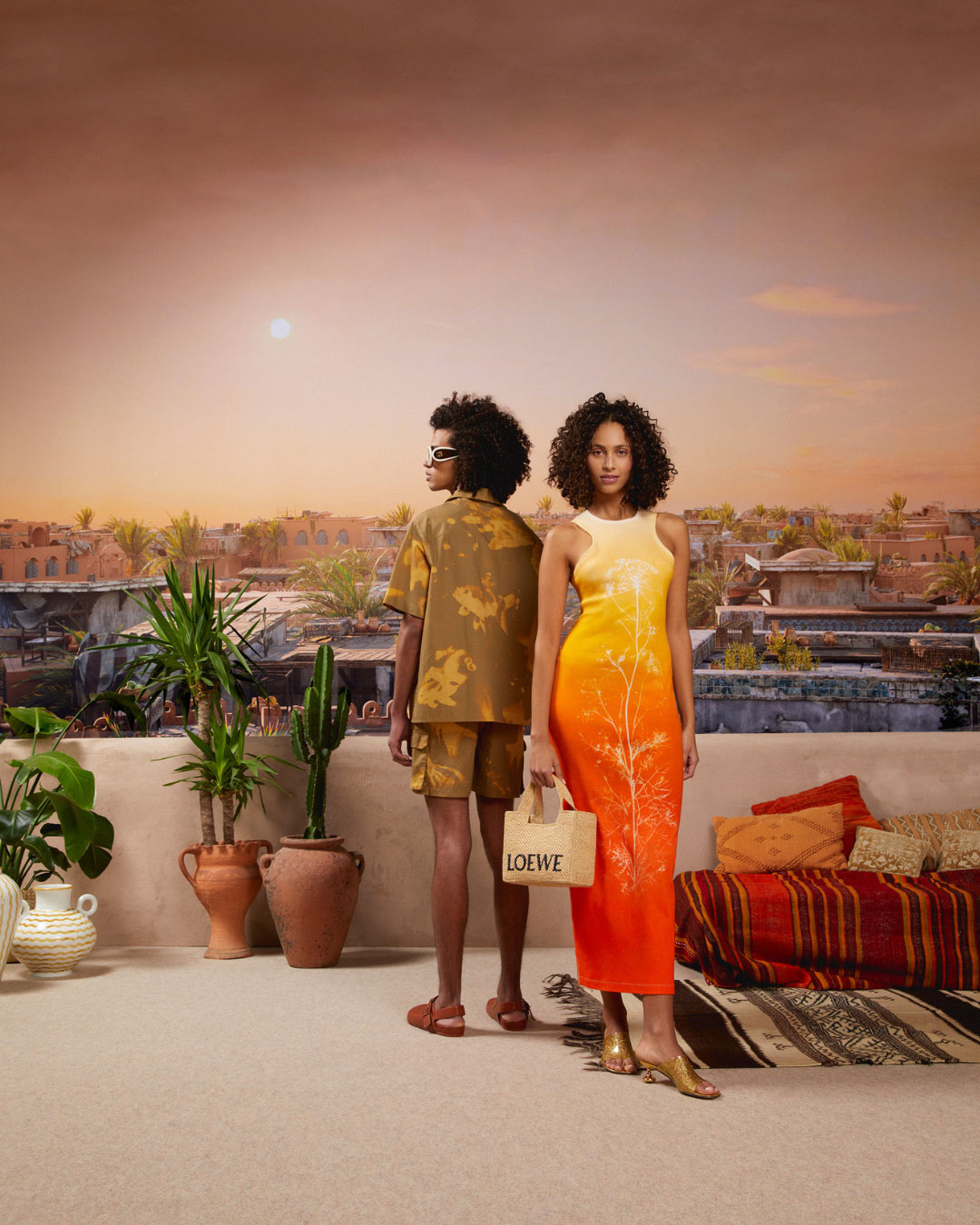 Deux mannequins posent sur une terrasse décorée de plantes et de jarres en terre cuite, avec un paysage de palmiers et de maisons en arrière-plan sous un ciel orangé. La femme porte une robe longue dégradée du jaune au orange et tient un sac Loewe, l’homme est en tenue estivale imprimée, lunettes blanches et sandales.