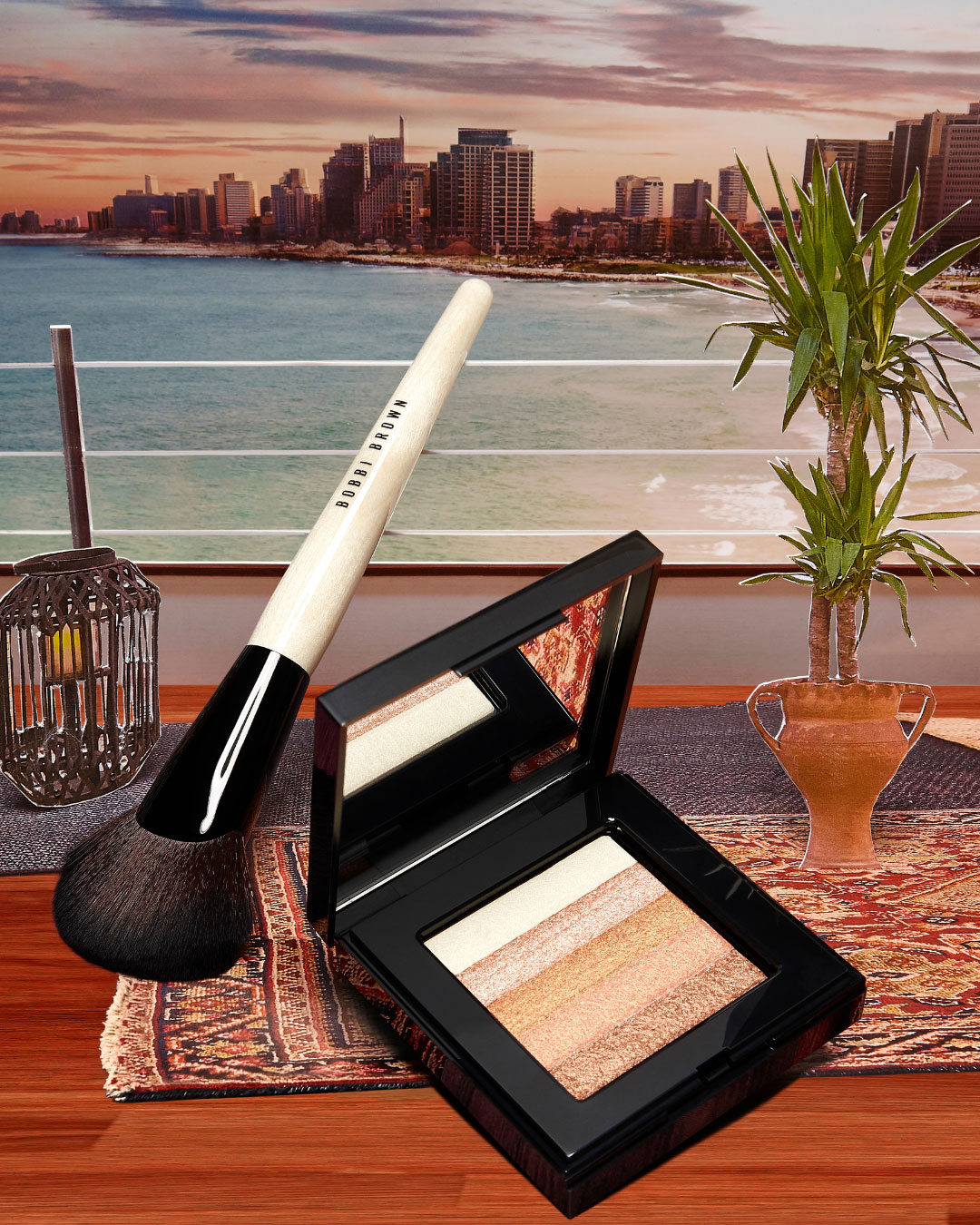 Un pinceau de maquillage repose sur une table en bois à côté d'une palette d’highlighter Bobbi Brown ouverte. L’arrière-plan montre une ville côtière moderne au coucher du soleil, vue depuis une terrasse décorée de plantes et d’un tapis oriental.