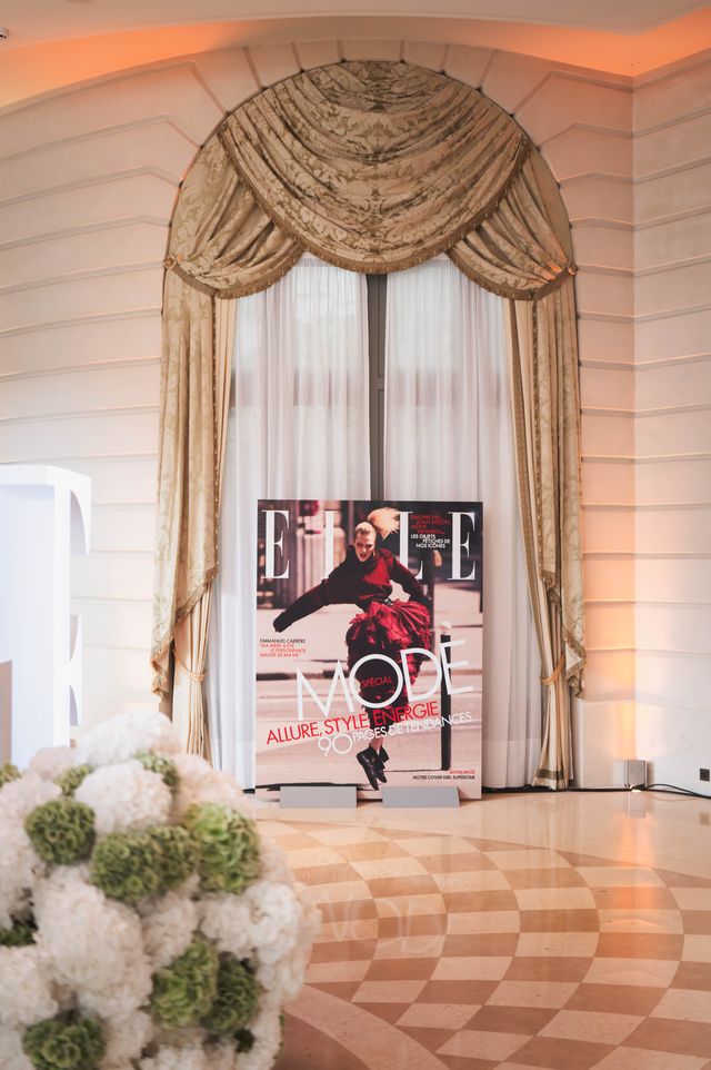 Affiche géante de la Une "Spécial Mode" du magazine ELLE installée à l’entrée d’un salon élégant, dans un décor raffiné avec rideaux drapés et lustres. La couverture met en vedette un mannequin souriant vêtu d’un manteau en fausse fourrure, avec le titre « Puissante, amoureuse, stylée » sur fond noir.