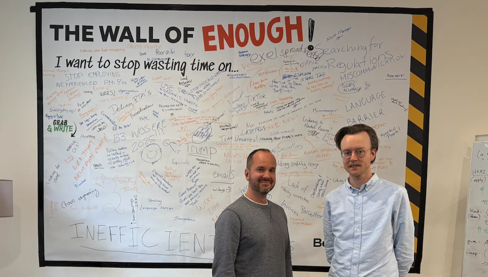 Dos hombres de pie frente a una pizarra cubierta de notas manuscritas titulada «The Wall of Enough»