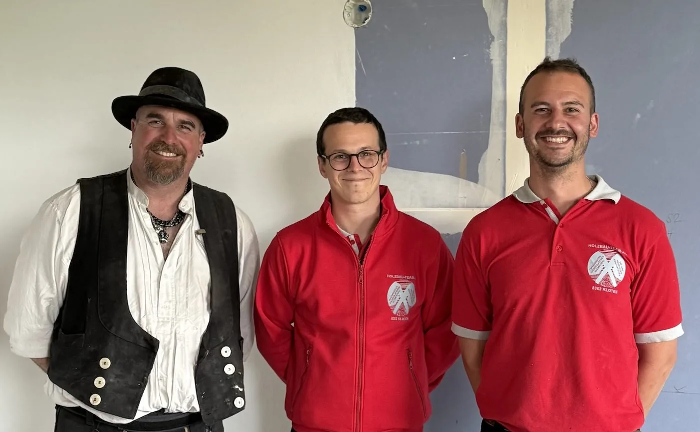 Drei Männer im Innenraum, einer in historischem Kostüm, zwei im roten Holzbau-Team-Poloshirt
