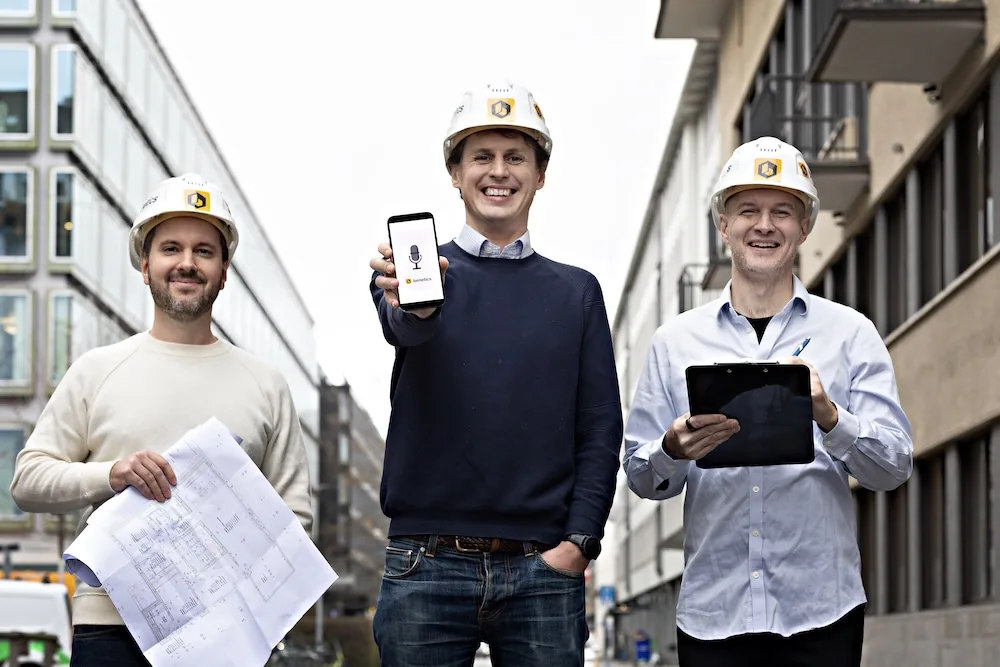 Three men in hard hats on a city street, one holding a phone with the Benetics app, one holding blueprints
