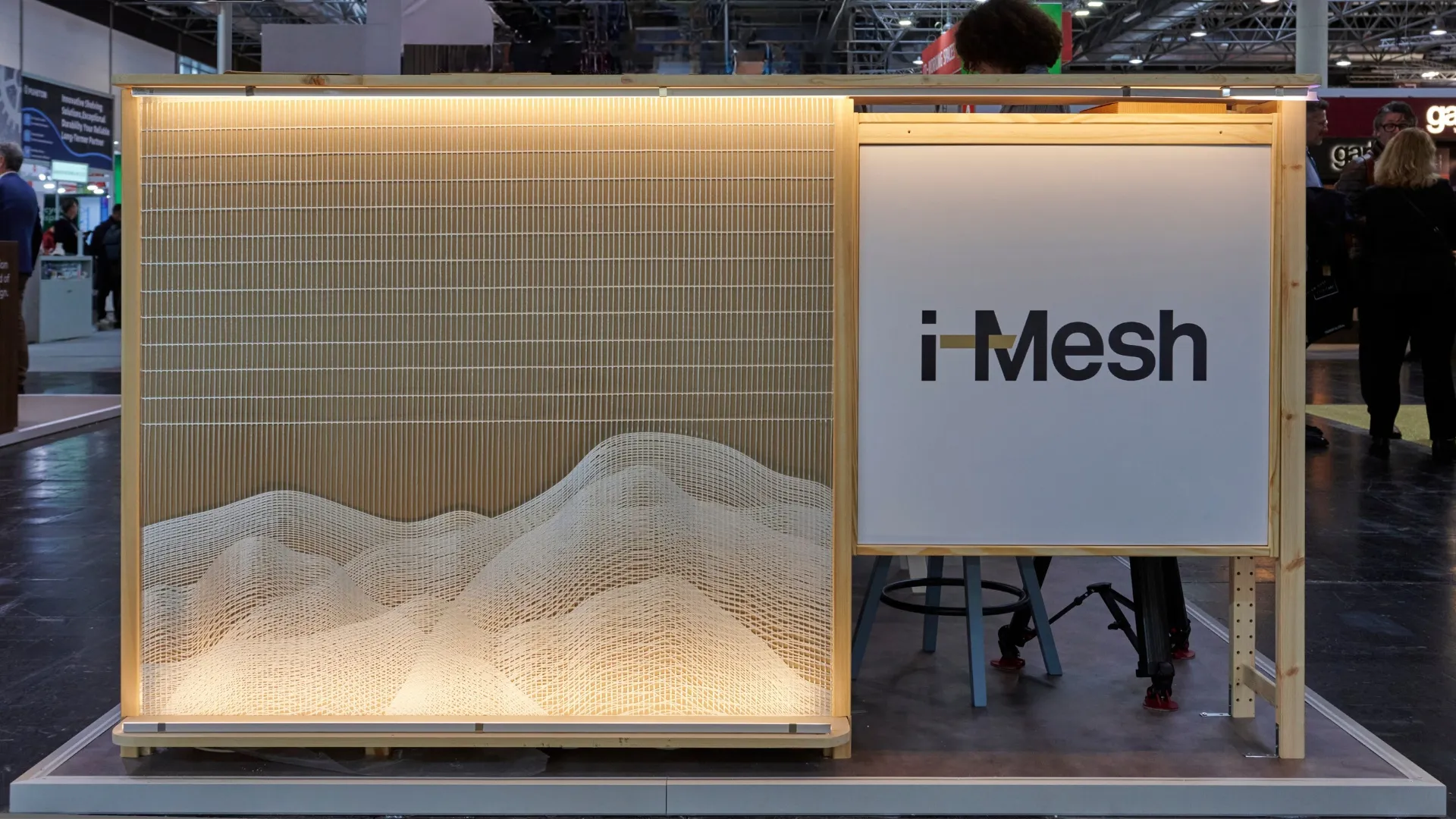 i-Mesh stand at Euroshop 2026 featuring a backlit wave-shaped architectural mesh installation in a wooden frame
