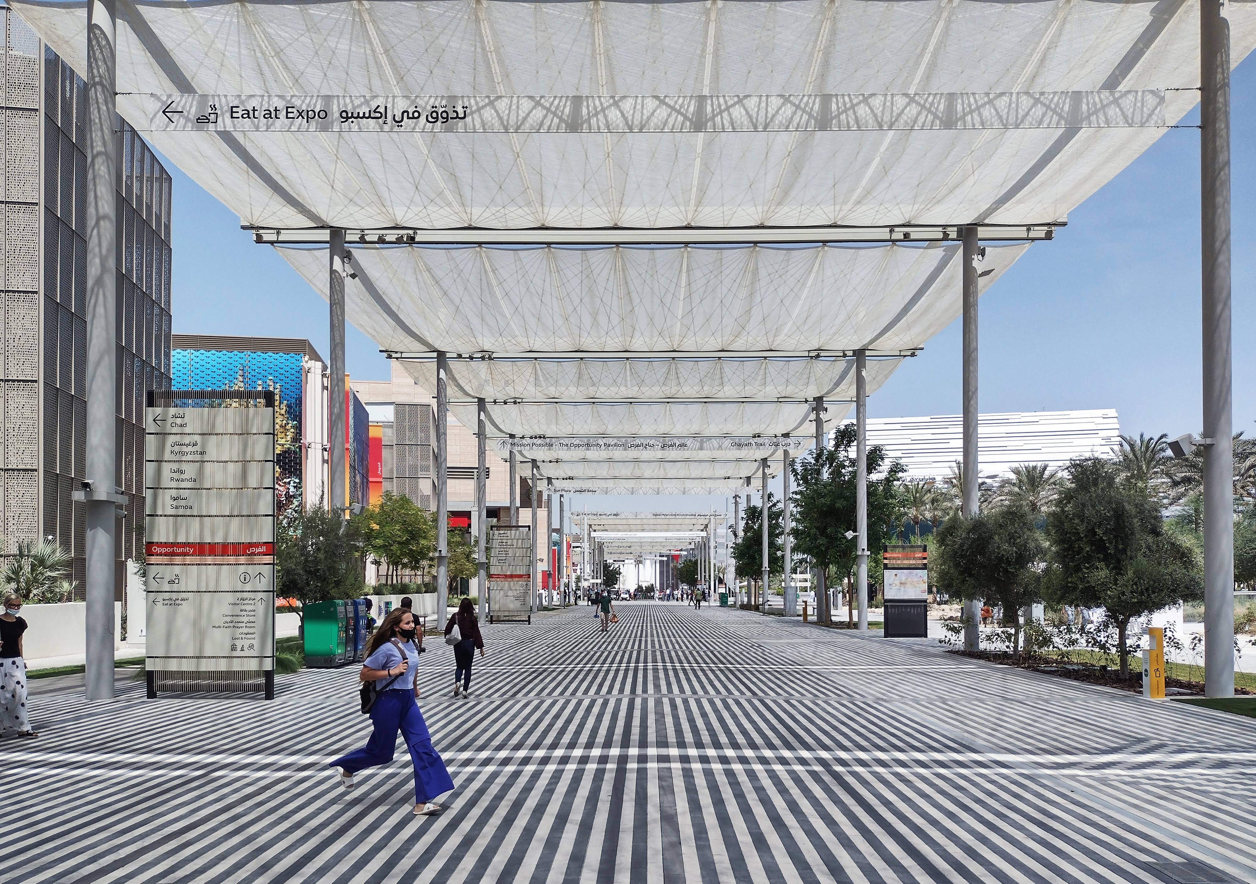 Perspective view of the Opportunity Concourse at Expo 2020 Dubai fully covered by the i-Mesh retractable solar shading system, with visitors walking along the striped pedestrian boulevard and national pavilions on both sides