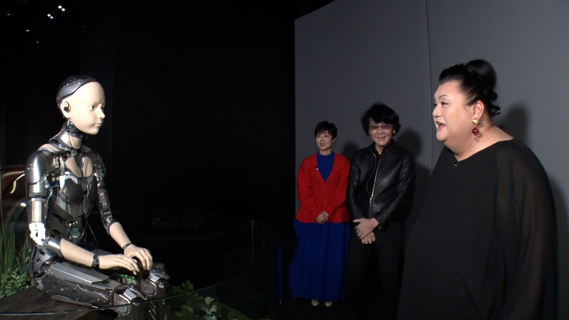 Tre visitatori interagiscono con un robot umanoide seduto dal volto realistico all'interno del padiglione Future of Life all'Expo 2025 di Osaka, in un ambiente poco illuminato con piante sullo sfondo