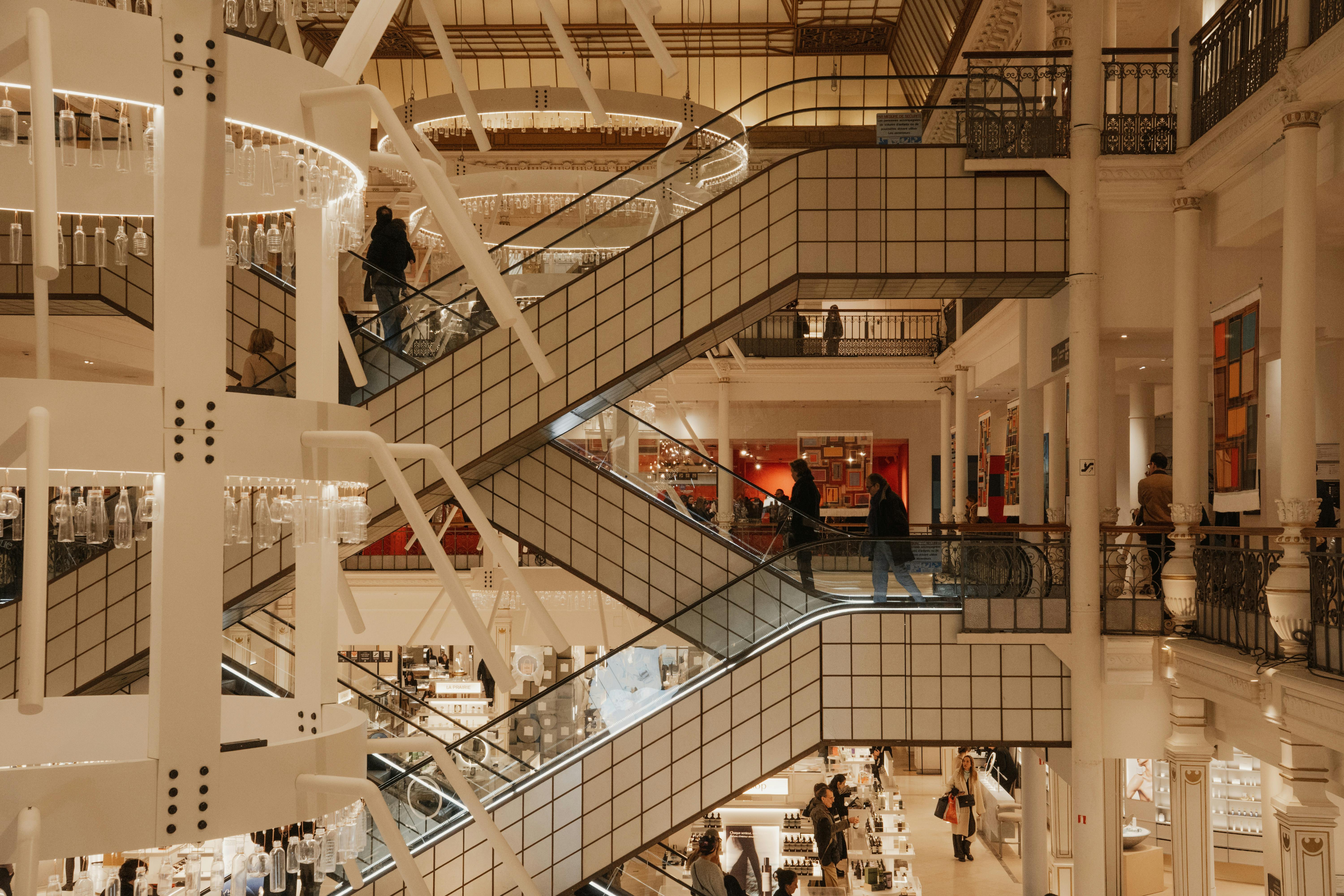 Interior of Le Bon Marché department store in Paris, showing multi-level escalators, white steel structure, ornate cast-iron columns and warm ambient lighting