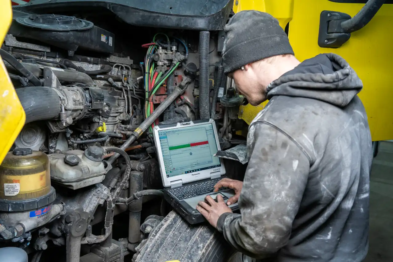 Technician runs diagnostics on a diesel engine using a rugged laptop with green and red status bars on screen. Ask ChatGPT