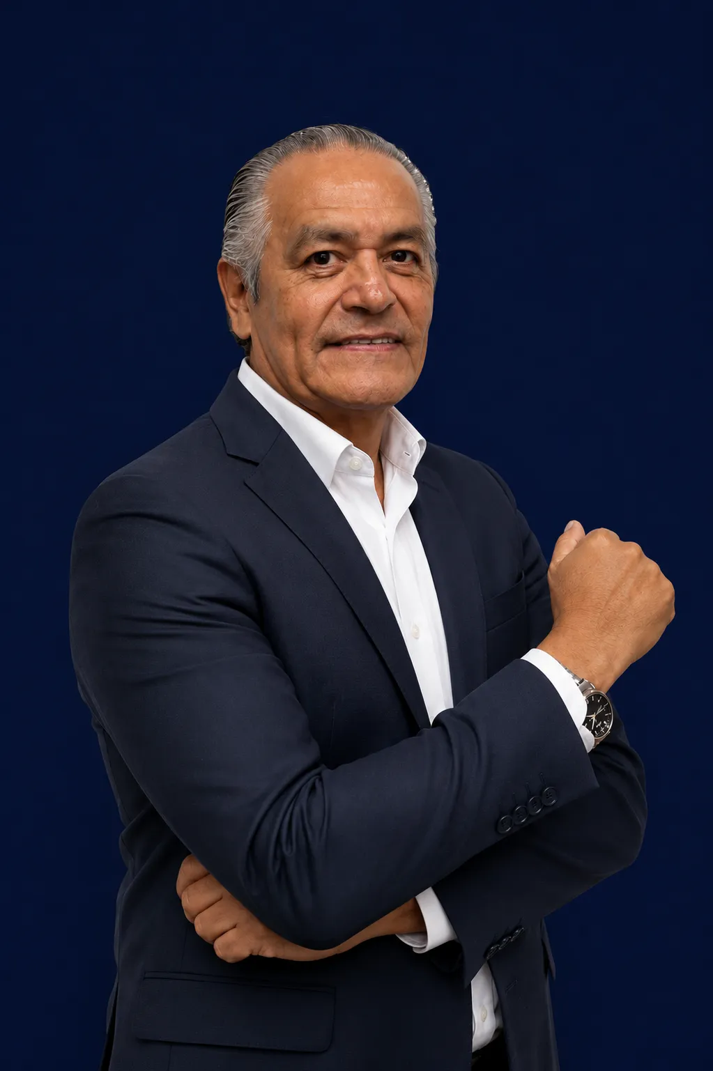 Hombre mayor con traje azul oscuro y camisa blanca, brazos cruzados, fondo azul.