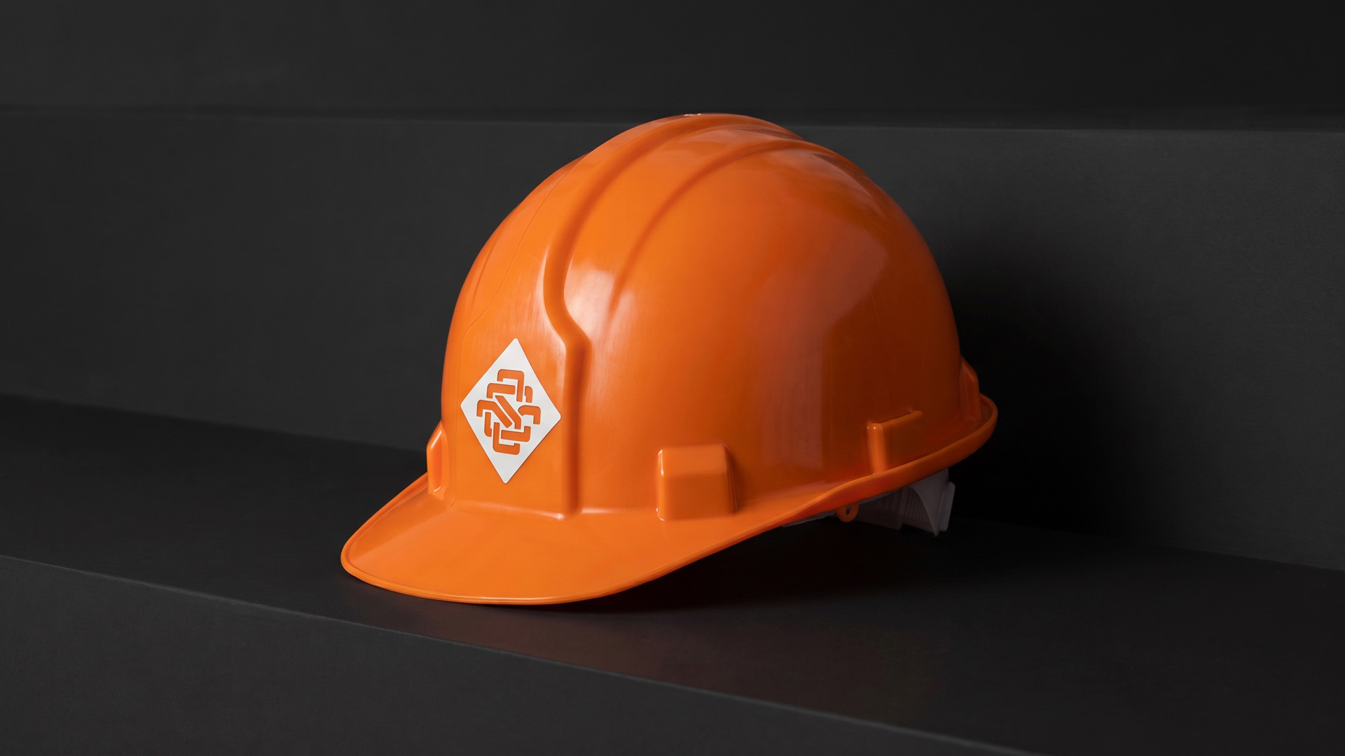 Orange hard hat with a white diamond logo badge, resting on a black stepped surface against a dark wall.