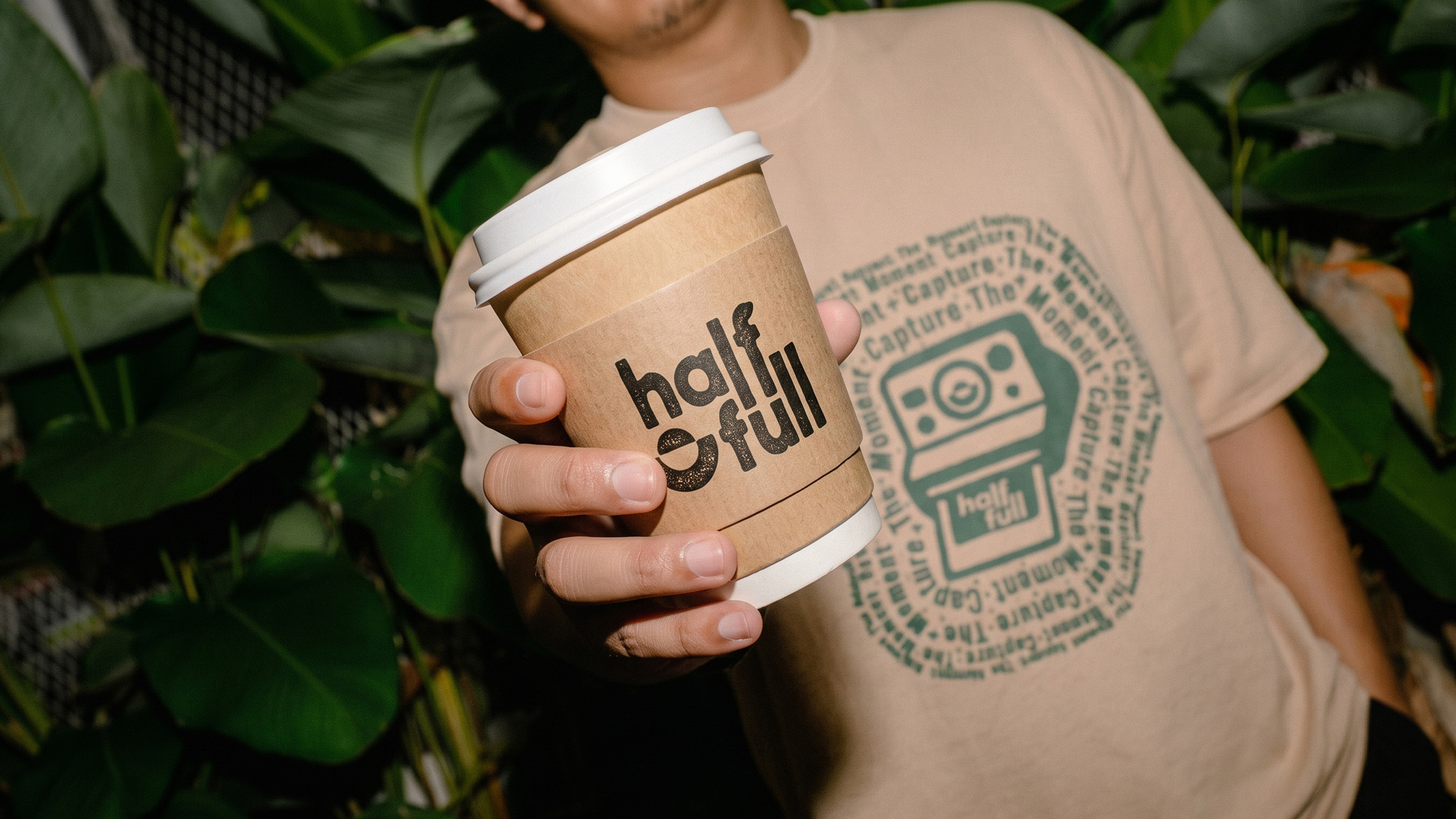 Hand holding a coffee cup with a kraft half full sleeve, in front of a tan tee printed with a green camera graphic.