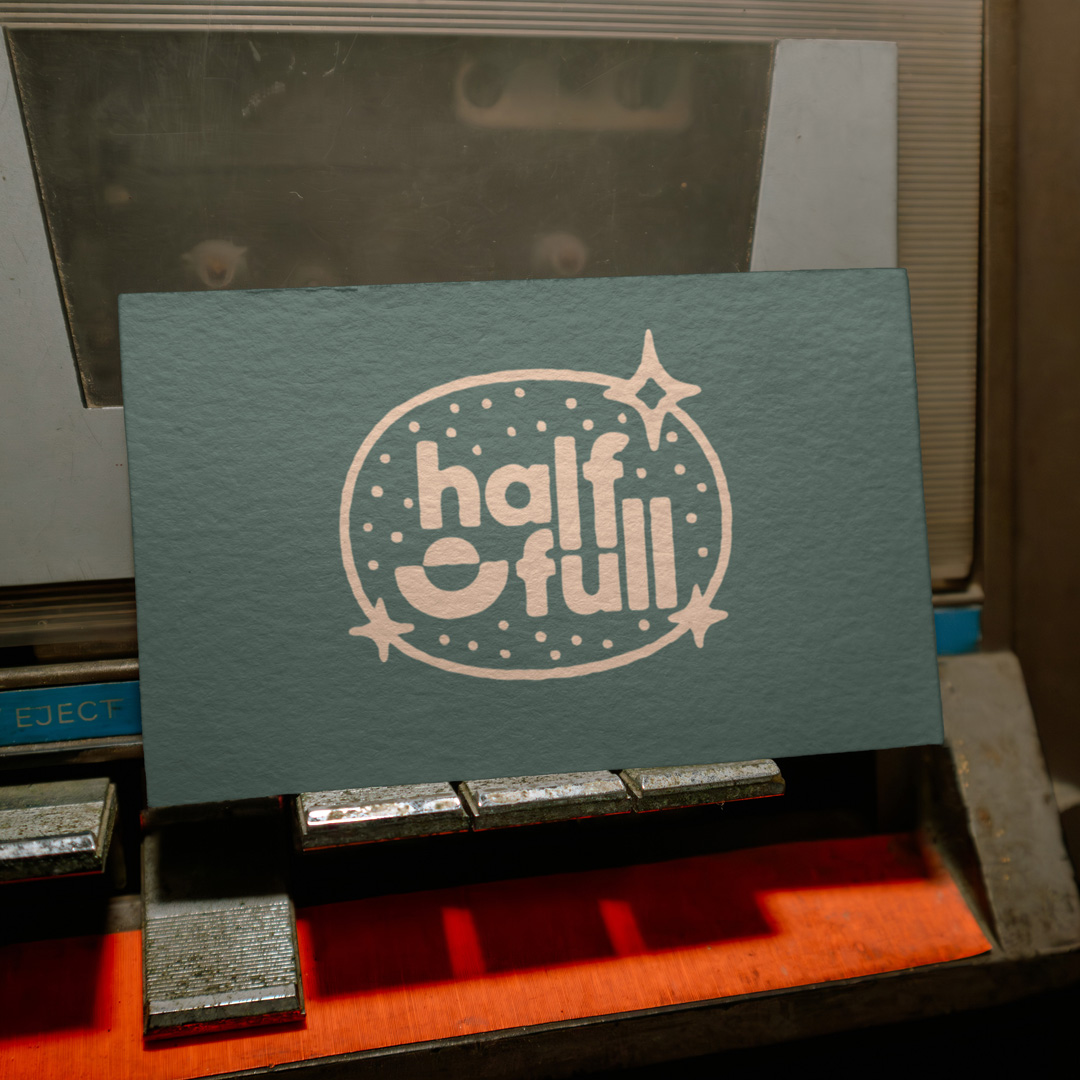 Green business card with a cream half full logo in a starred oval, propped against an old cassette deck.