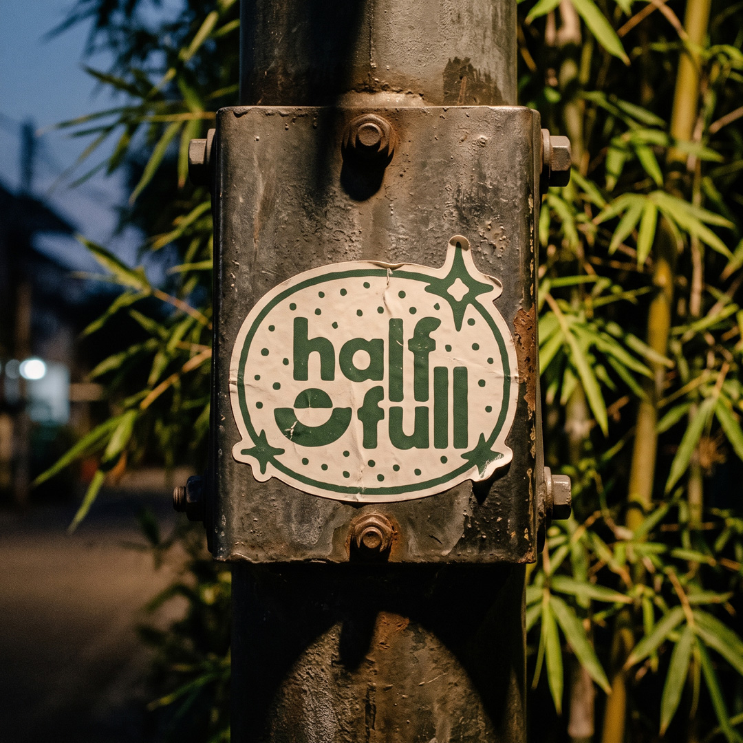 Cream half full sticker with a green logo, stuck to a weathered metal pole in front of bamboo.