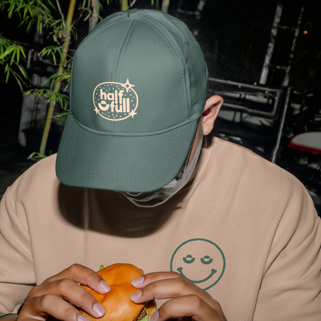 Person in a green cap with a cream half full logo and a tan sweater with a green smiley face, holding a burger at night.