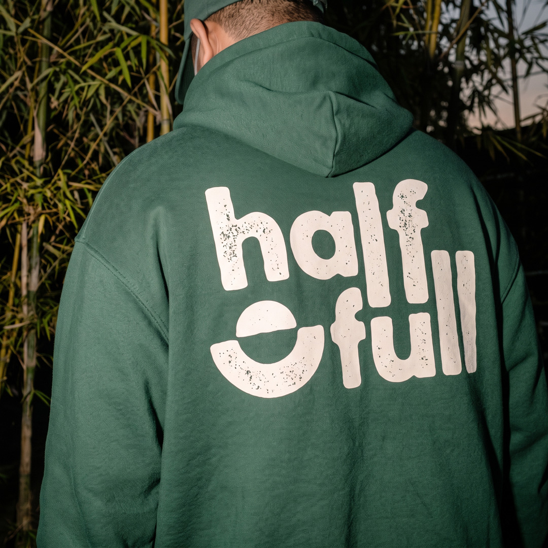 Back of a green hoodie with a large cream half full wordmark, photographed in front of bamboo.