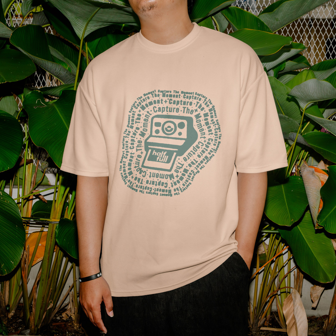 Person in a tan tee with a green camera and Capture the Moment graphic, standing in front of large leaves.